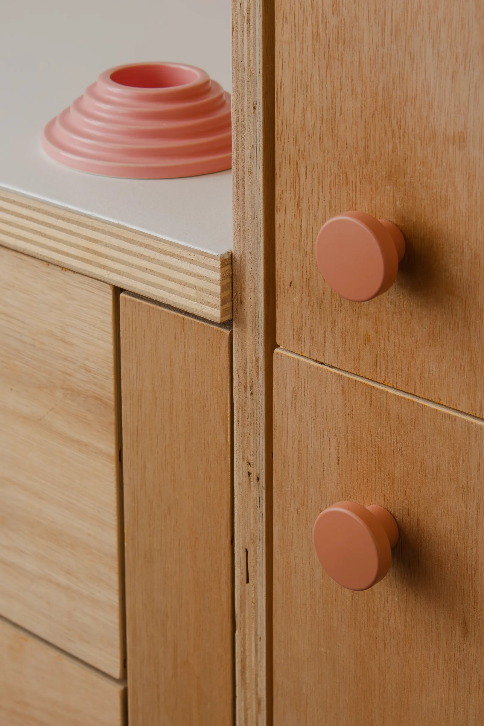 Gros plan sur des armoires de cuisine en bois avec des poignées rondes couleur terre cuite et un plan de travail blanc sur lequel repose un coquetier rose Axel Chay.