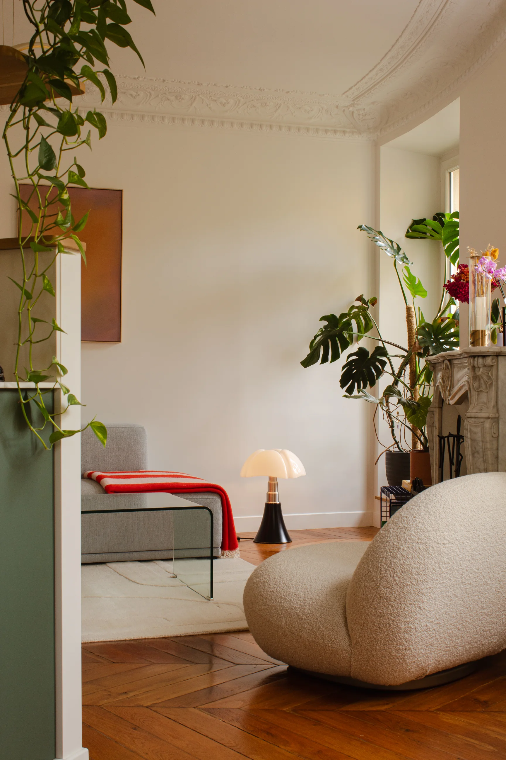 Coin salon confortable avec un fauteuil moderne beige aux lignes arrondies, une table basse en verre, un canapé gris recouvert d'un plaid à rayures rouges, des plantes décoratives et des moulures ornées au plafond.