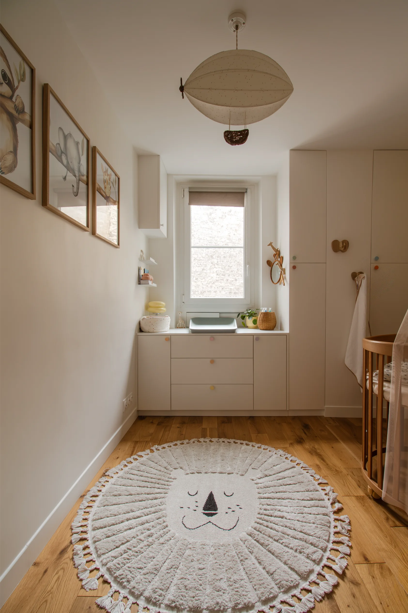 Chambre d'enfant aux tons neutres avec parquet, tapis rond en forme de tête de lion, armoires blanches sous la fenêtre, décorations murales représentant des animaux et lampe suspendue en forme de montgolfière.