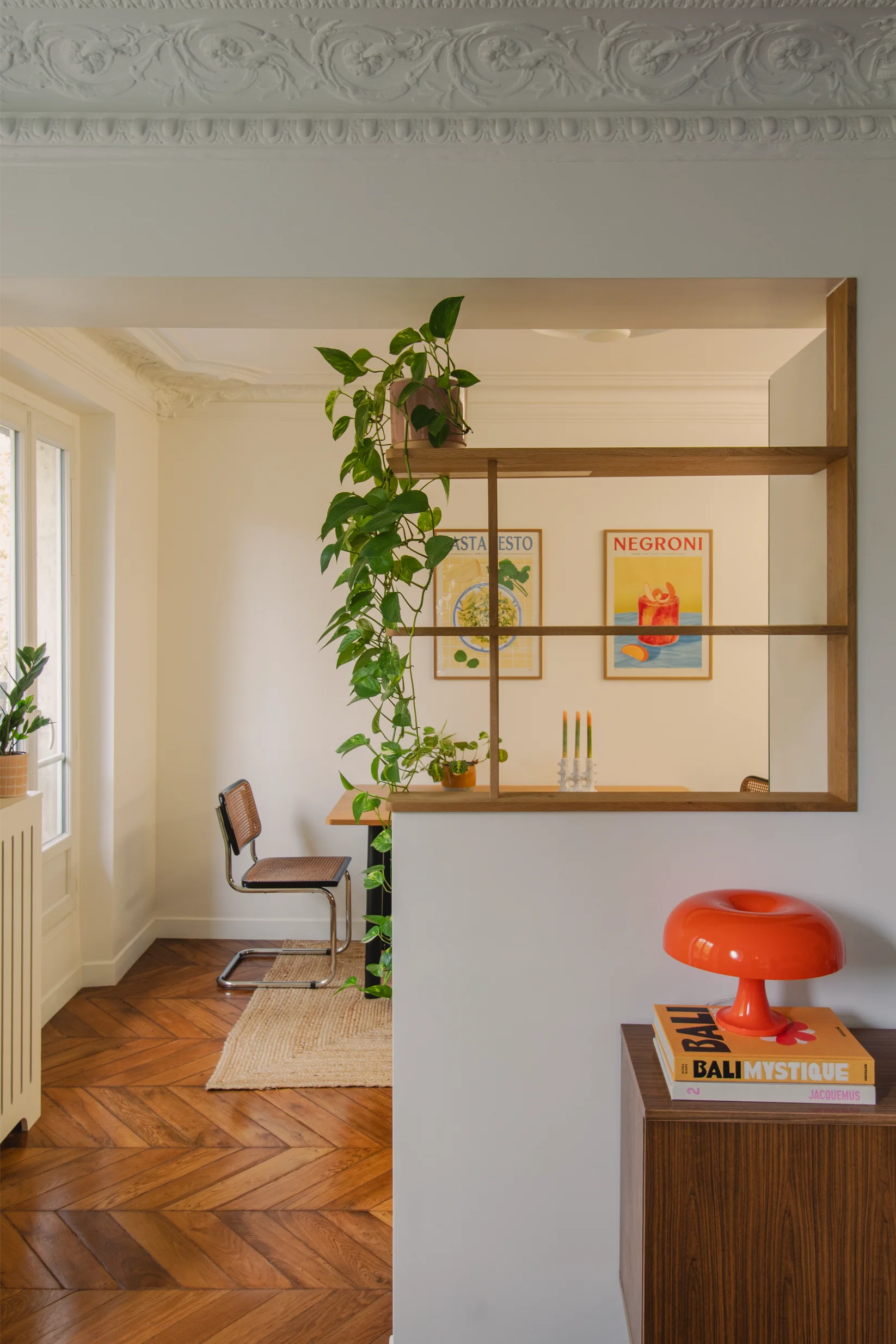 Salle à manger lumineuse avec parquet à chevrons, chaise du milieu du siècle, décor mural représentant des pâtes au pesto et un negroni, plantes vertes et lampe orange moderne Nesso Artemide posée sur un meuble en bois.