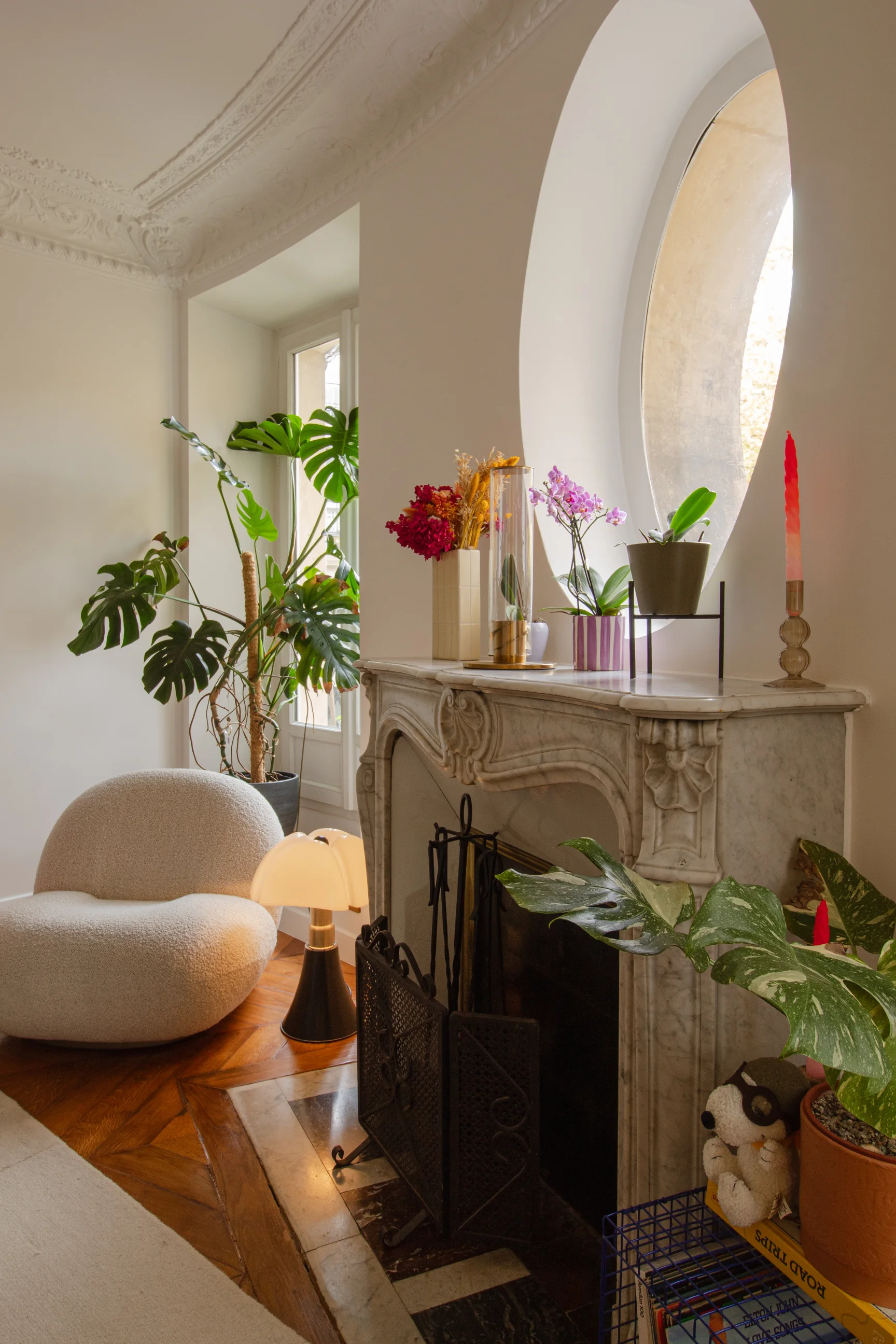 Coin salon confortable avec un fauteuil beige moelleux, une lampe de table moderne, une cheminée en marbre ornée de plantes en pot et de fleurs, et une grande plante à feuilles près d'une grande fenêtre.