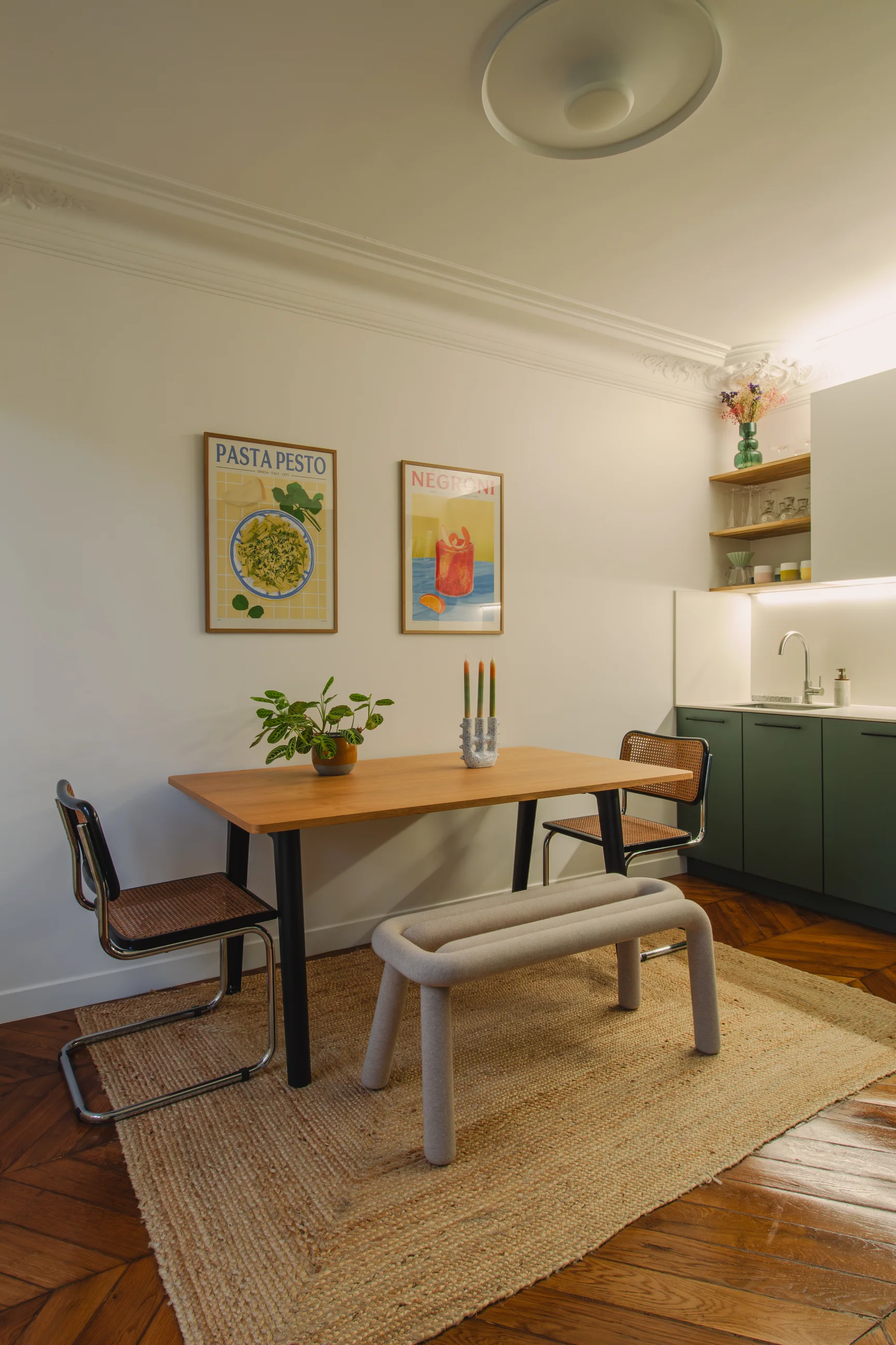 Coin repas confortable avec une table en bois, deux chaises en osier, un banc rembourré posé sur un tapis tissé et des affiches encadrées représentant des plats et des boissons accrochées au mur.
