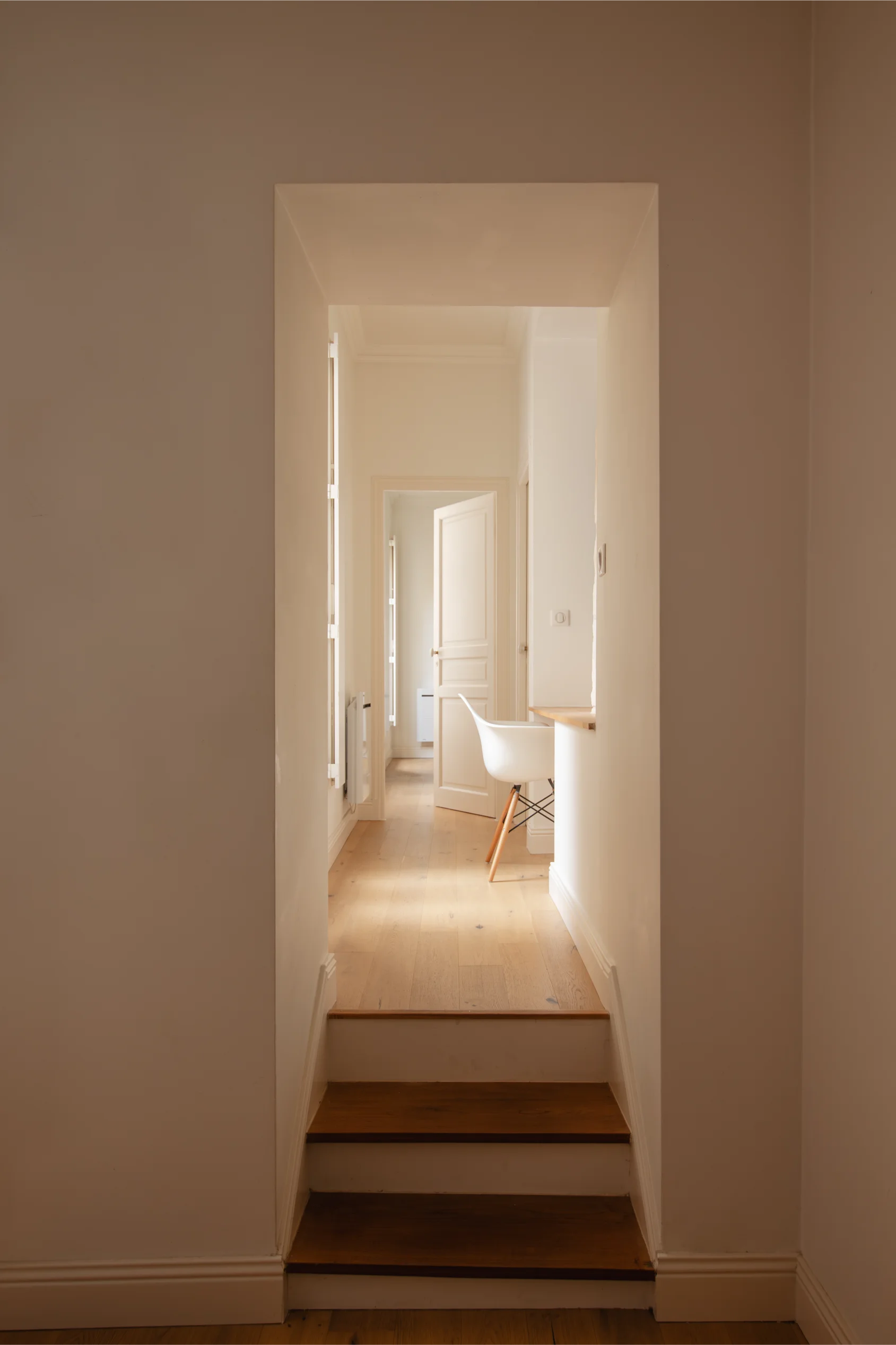 Couloir étroit avec trois marches en bois menant à une pièce lumineuse avec un sol en bois clair, des murs blancs, une chaise blanche et une porte ouverte.
