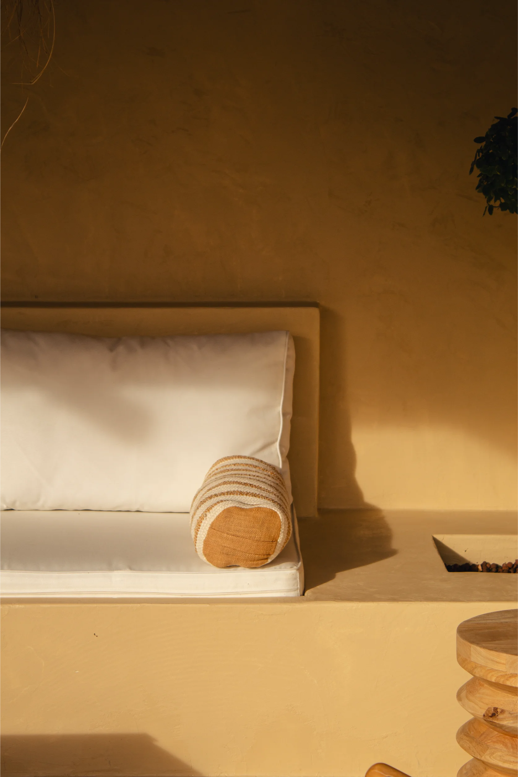 Gros plan sur un banc rembourré blanc avec un coussin cylindrique rayé beige et blanc, adossé à un mur texturé couleur beige chaud, à côté d'une table ronde en bois.