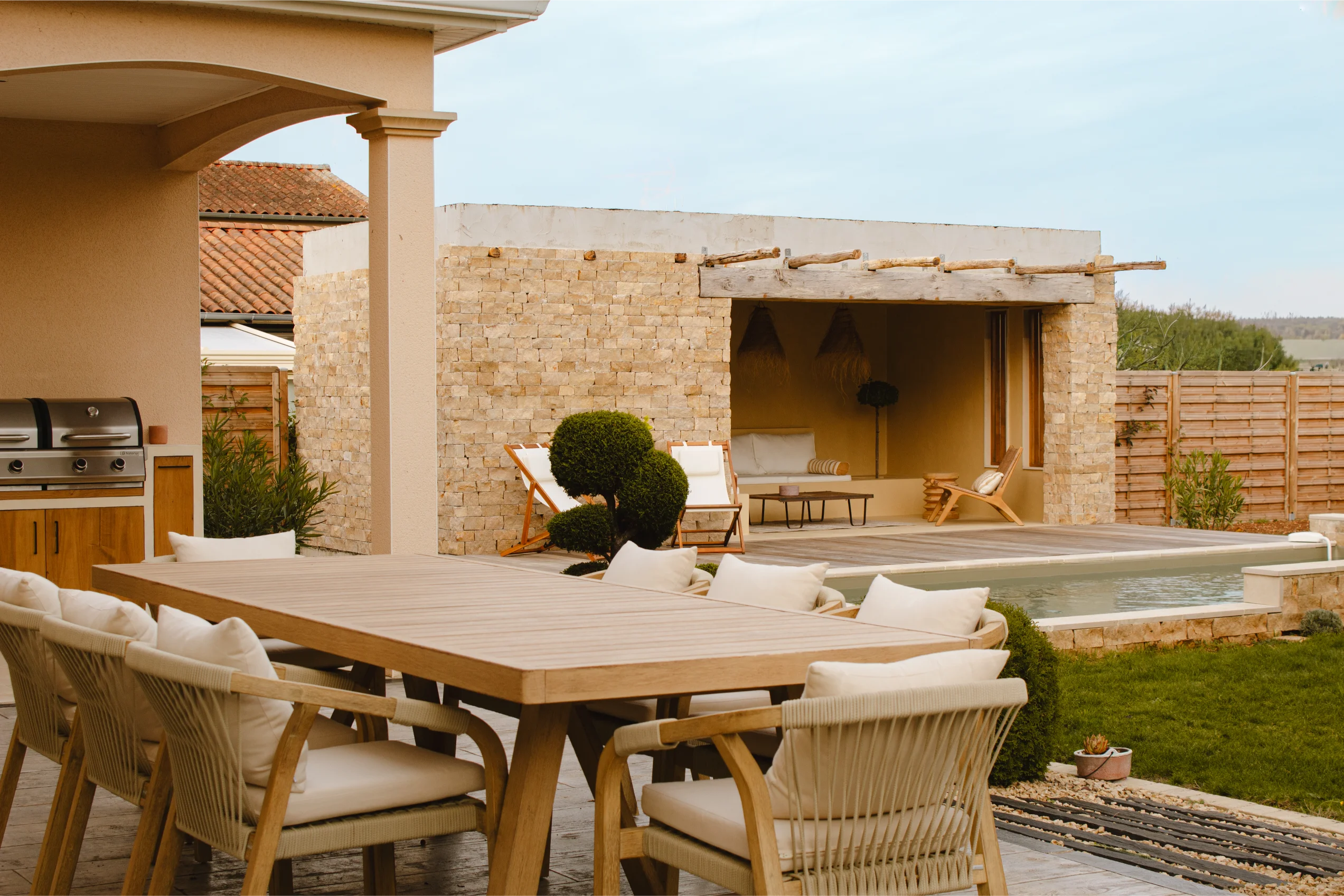 Coin repas extérieur avec table en bois et chaises rembourrées à côté d'un pool house en pierre et d'une petite piscine.
