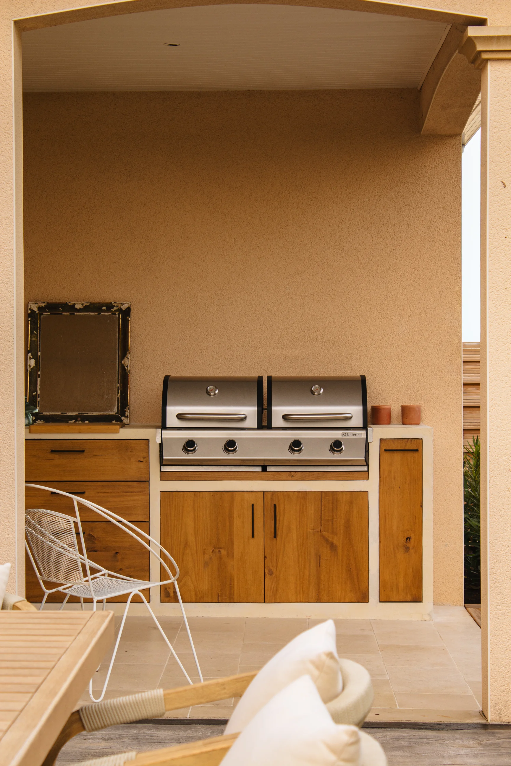 Cuisine extérieure avec double grill en acier inoxydable, armoires en bois et chaise en métal blanc.