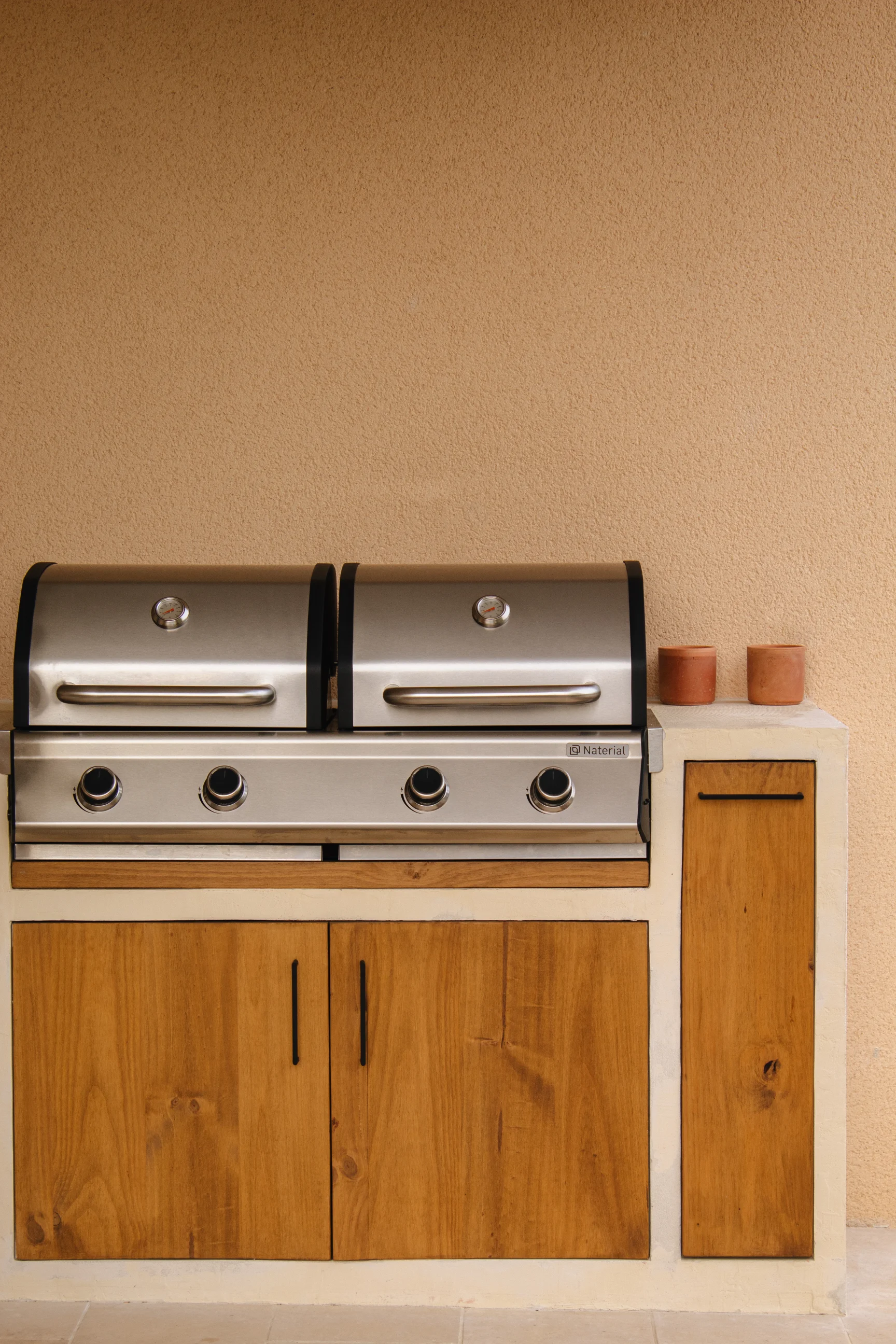 Barbecue extérieur moderne en acier inoxydable avec deux couvercles et quatre boutons de commande, encastré dans un meuble en bois et plâtre, contre un mur beige texturé.