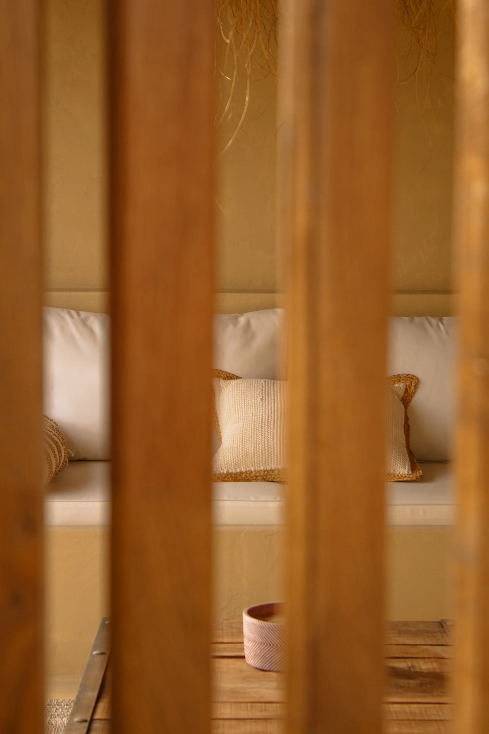 Canapé blanc rembourré avec des coussins beiges visible à travers des lattes de bois verticales, avec une table en bois et un petit bol devant.