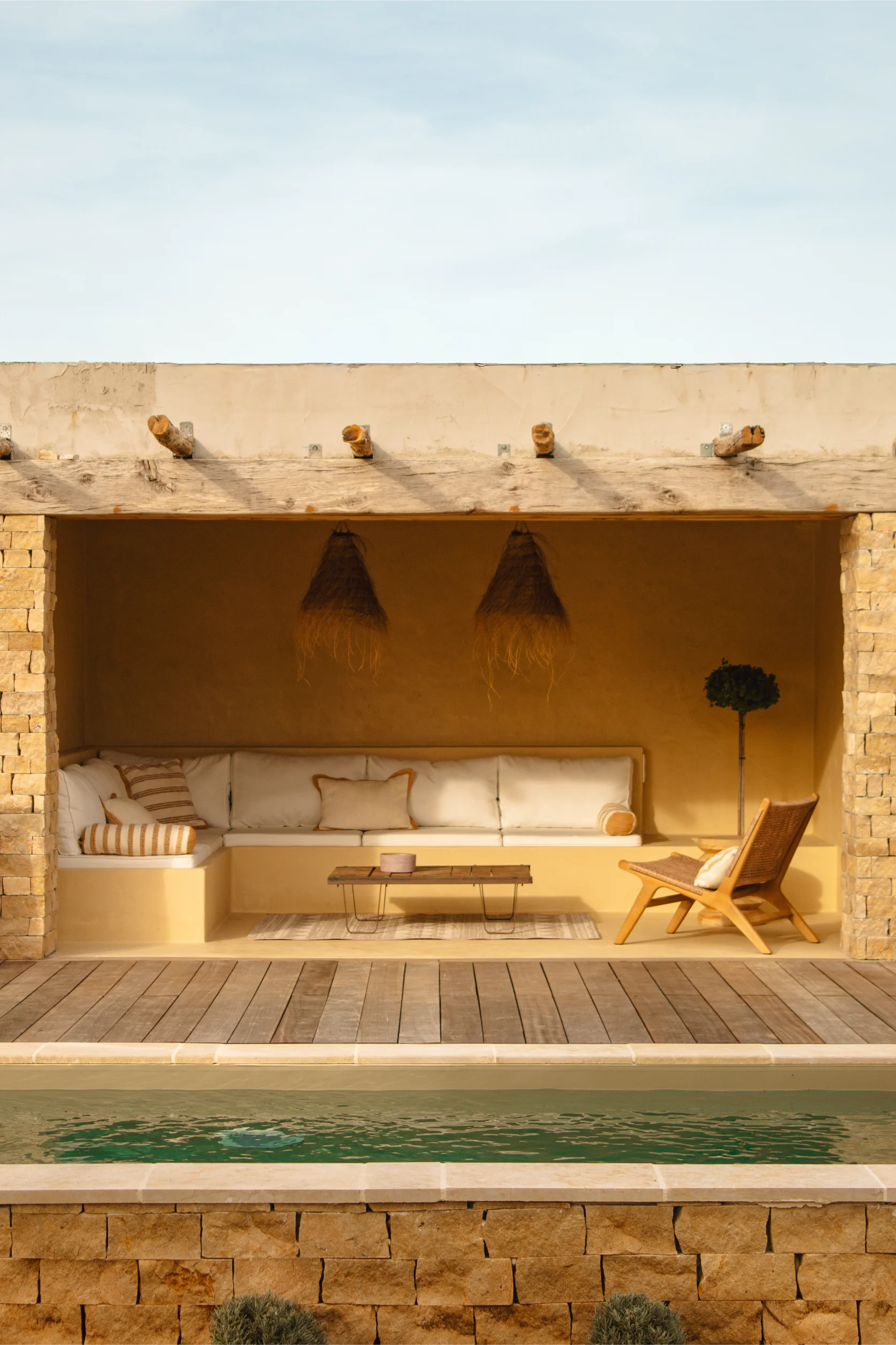 Coin salon extérieur confortable avec coussins blancs, chaise en bois et table sous un auvent beige à côté d'une piscine.
