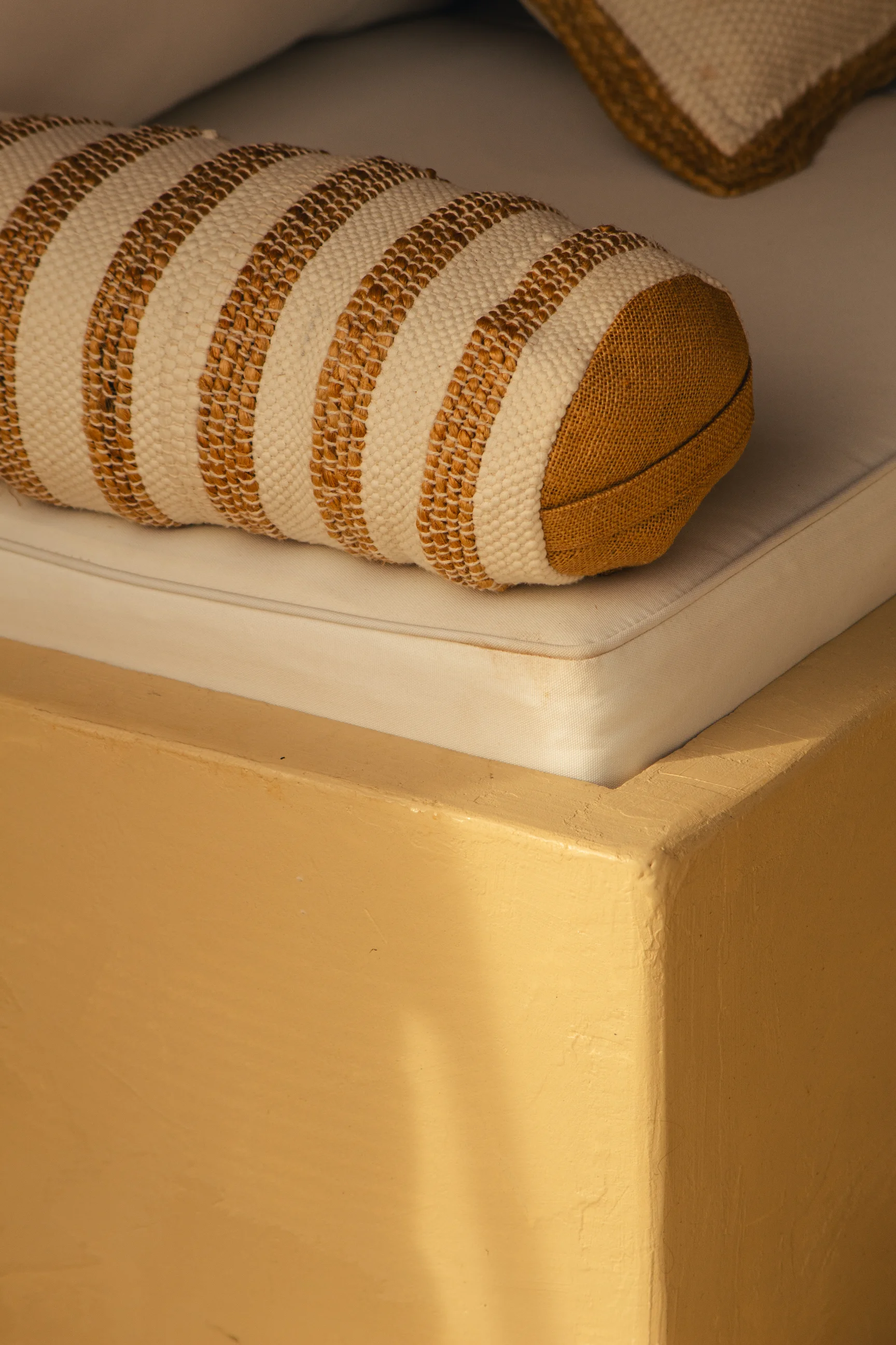 Gros plan sur un coussin rond rayé beige et moutarde posé sur un matelas blanc, avec un coin de mur jaune éclairé par la lumière du soleil.