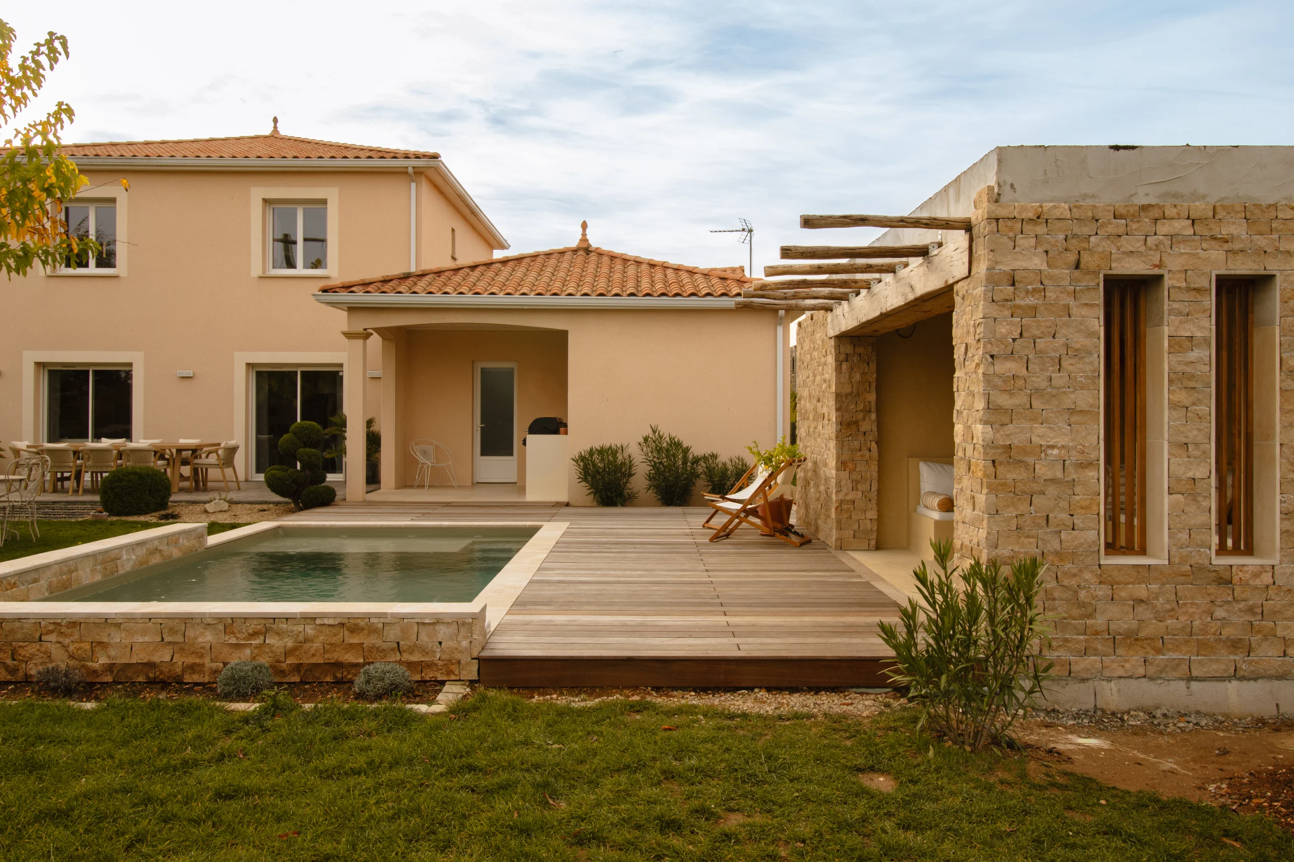 Arrière-cour avec une petite piscine, terrasse en bois avec chaises longues, maison beige et structure en pierre avec poutres en bois.