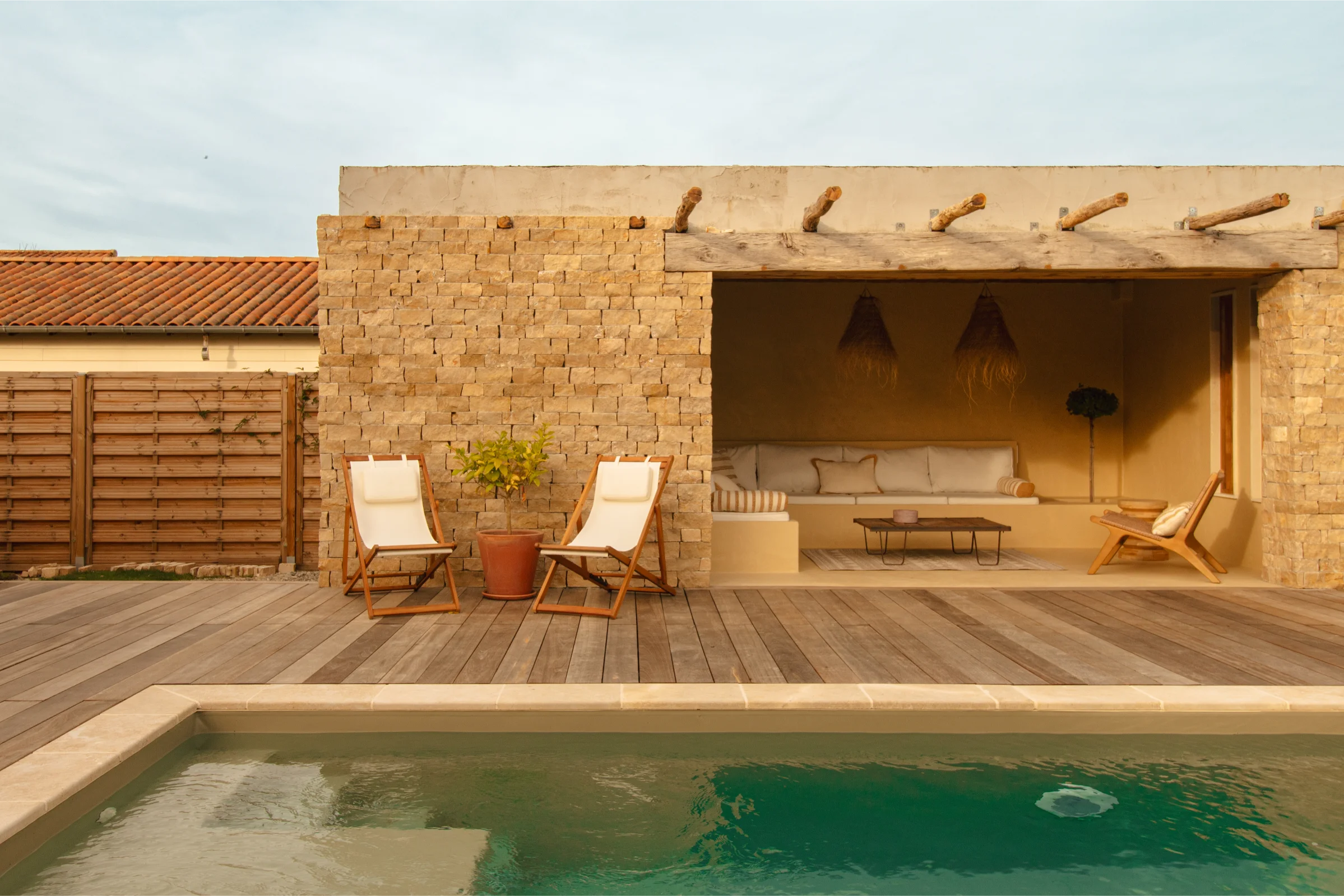 Terrasse moderne au bord de la piscine avec sol en bois, deux chaises longues avec coussins, plante en pot et coin salon ombragé avec canapé, table basse et suspensions en osier.