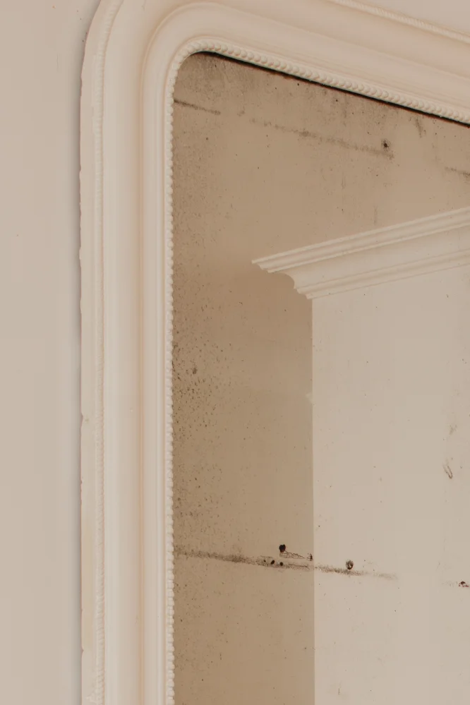 Gros plan d'un miroir vintage avec un cadre blanc orné, montrant des taches de vieillesse et un coin de moulure blanche reflétée.
