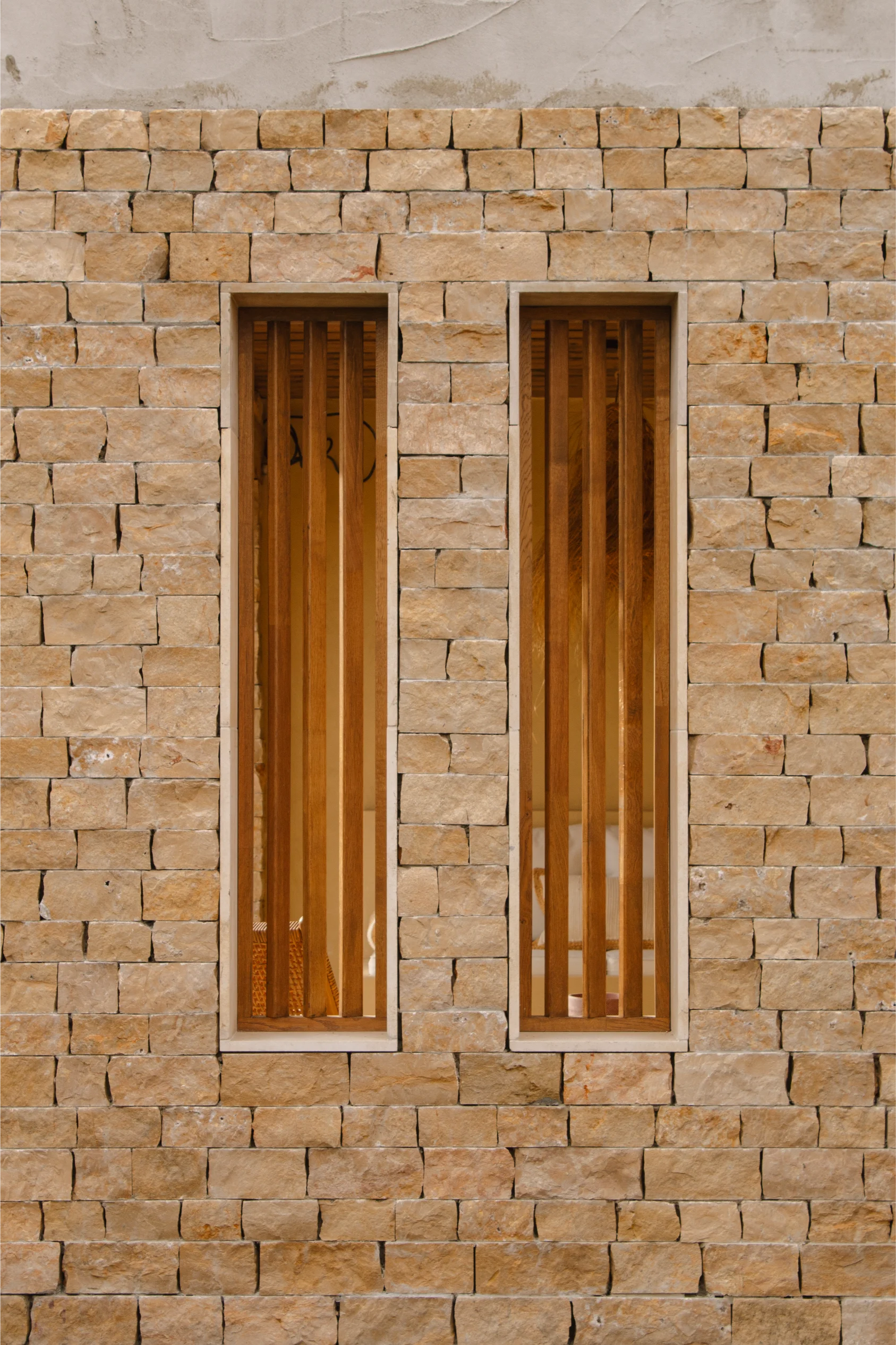Gros plan d'un mur en pierre beige avec deux fenêtres verticales étroites dotées de lattes en bois.