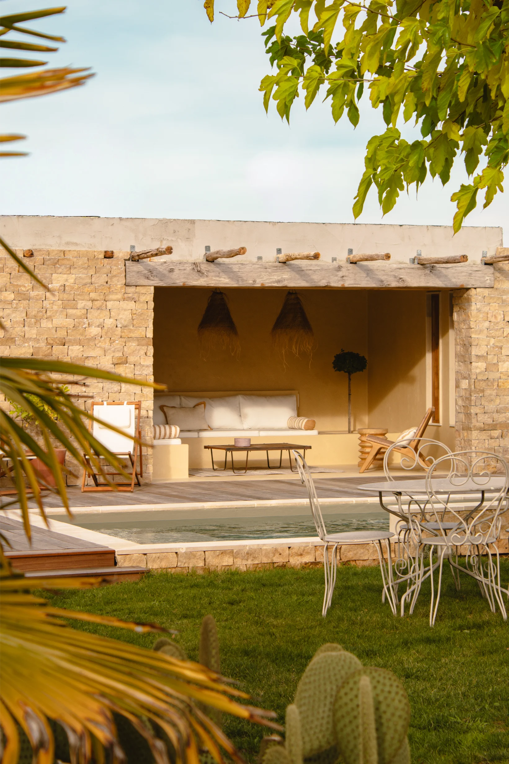 Coin salon extérieur confortable avec coussins blancs sous une pergola en bois, à côté d'une piscine et d'une pelouse verte, avec des chaises de jardin en métal.