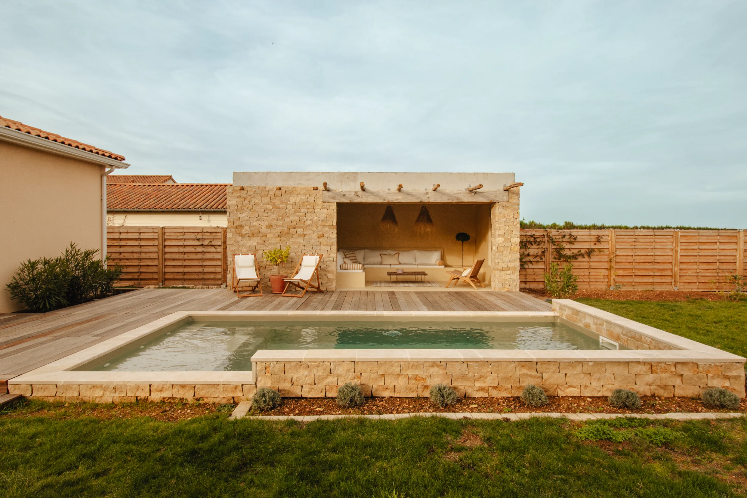 Jardin moderne avec piscine rectangulaire bordée de pierres, terrasse en bois et coin salon couvert avec chaises longues et coussins.