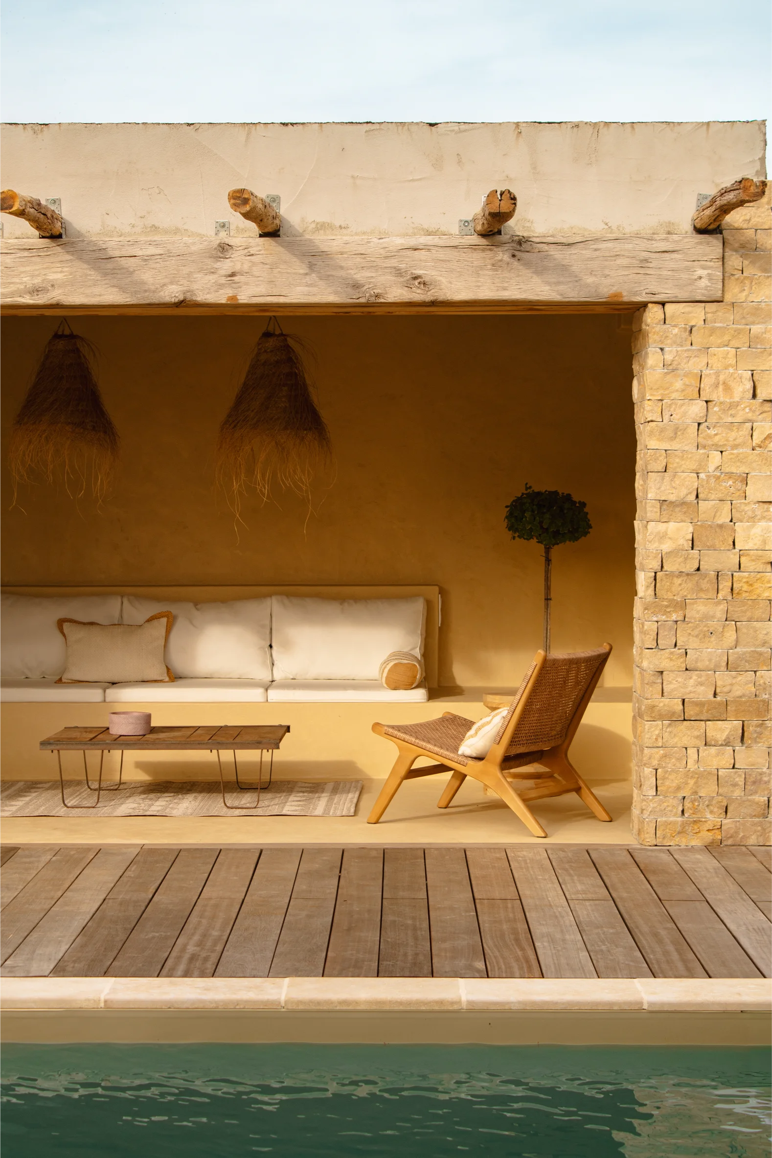 Espace salon extérieur près de la piscine avec chaise longue en bois, banc rembourré intégré, table basse en bois, lampes suspendues en fibres naturelles et mur en pierre.