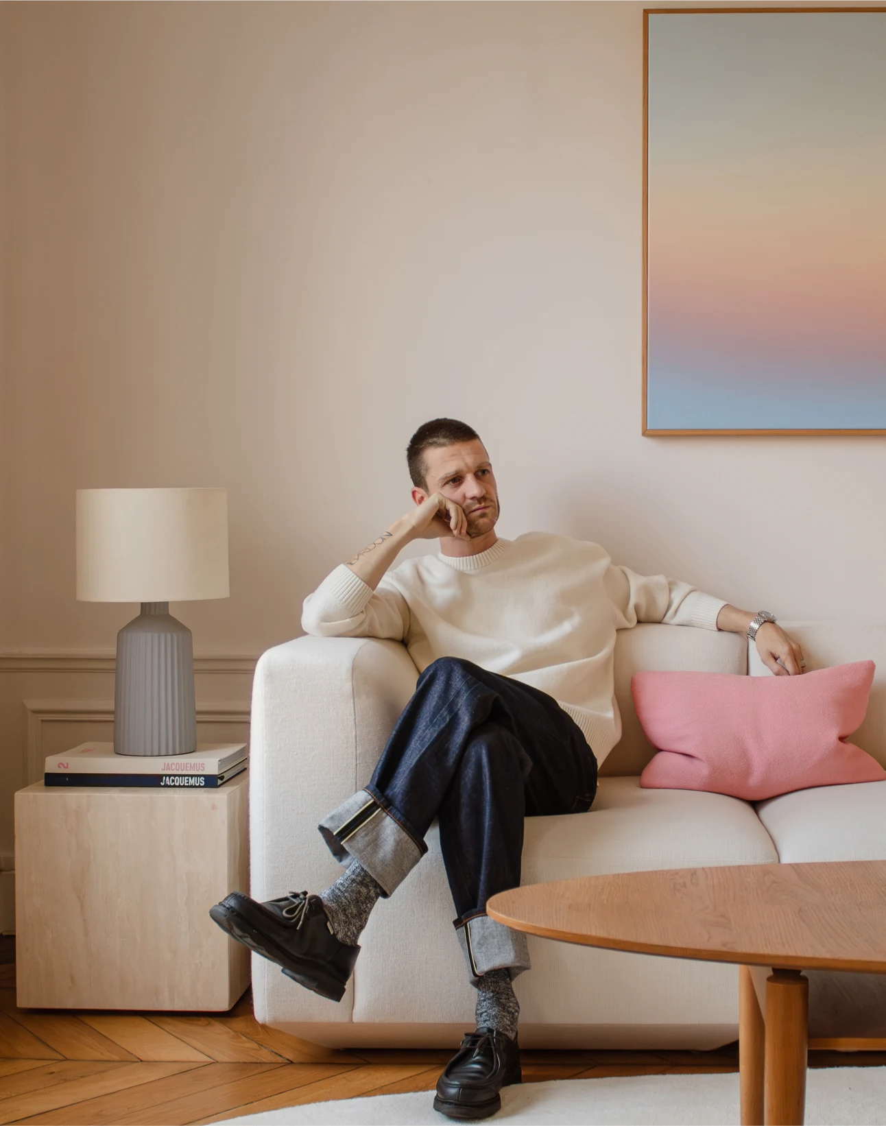Homme vêtu d'un pull blanc (Alexis Landrevie) et d'un jean retroussé, assis pensivement sur un canapé crème avec un coussin rose dans un salon minimaliste.