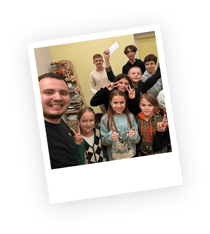 Group of smiling children and a young man posing indoors, some showing peace signs and thumbs up.