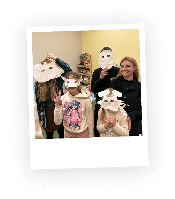 Group of five children indoors wearing and holding handmade white paper masks, one child making a peace sign and a girl without mask smiling.
