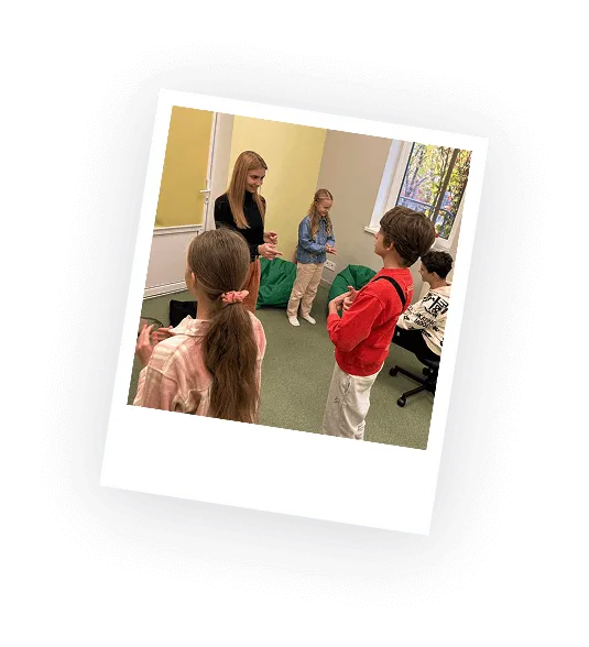 Group of children and a woman standing and engaging in an indoor activity in a room with green carpet and yellow walls.