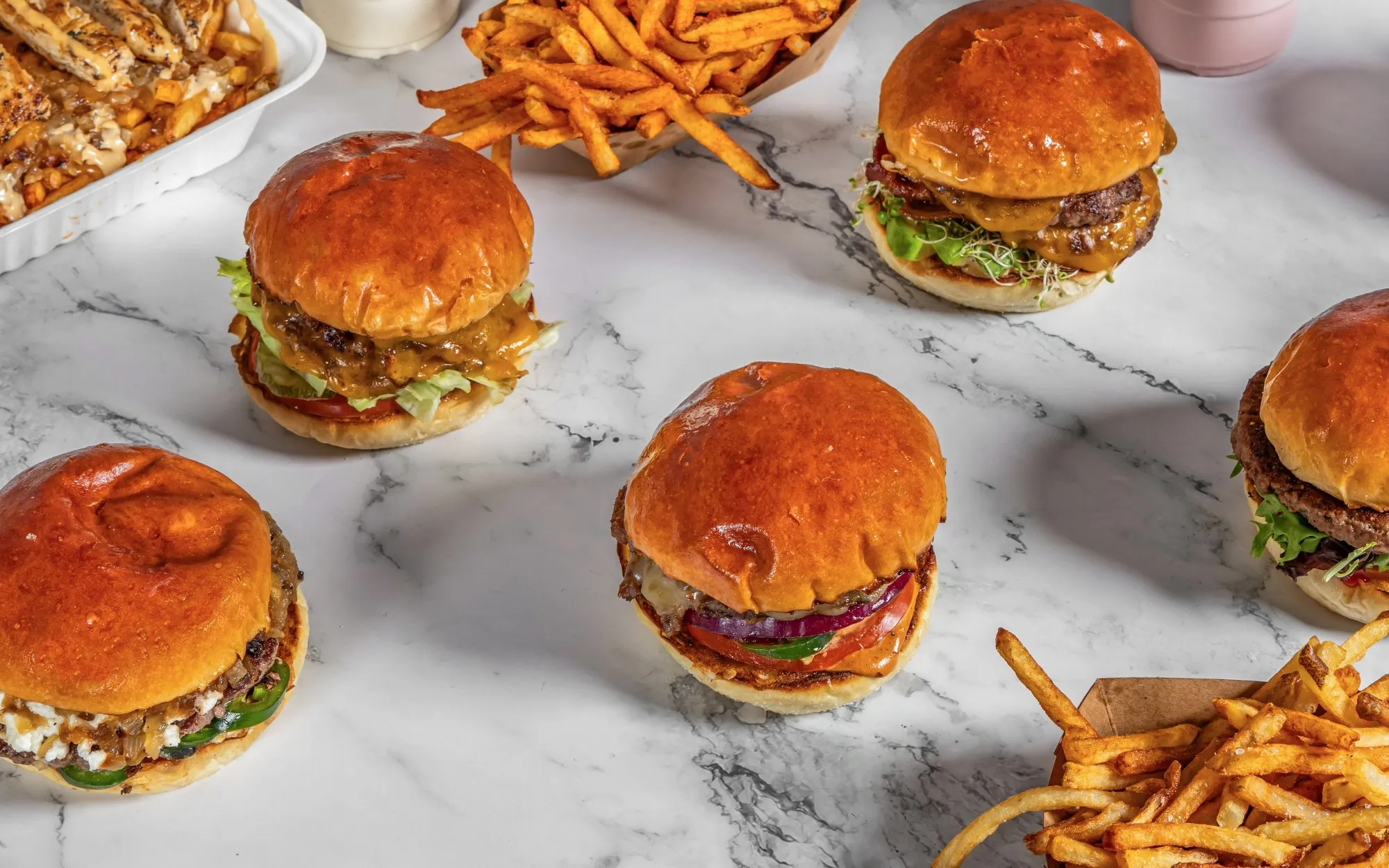 Aerial view of five assorted burgers with glossy buns on a marble table, surrounded by trays of crispy loaded fries and regular fries.
