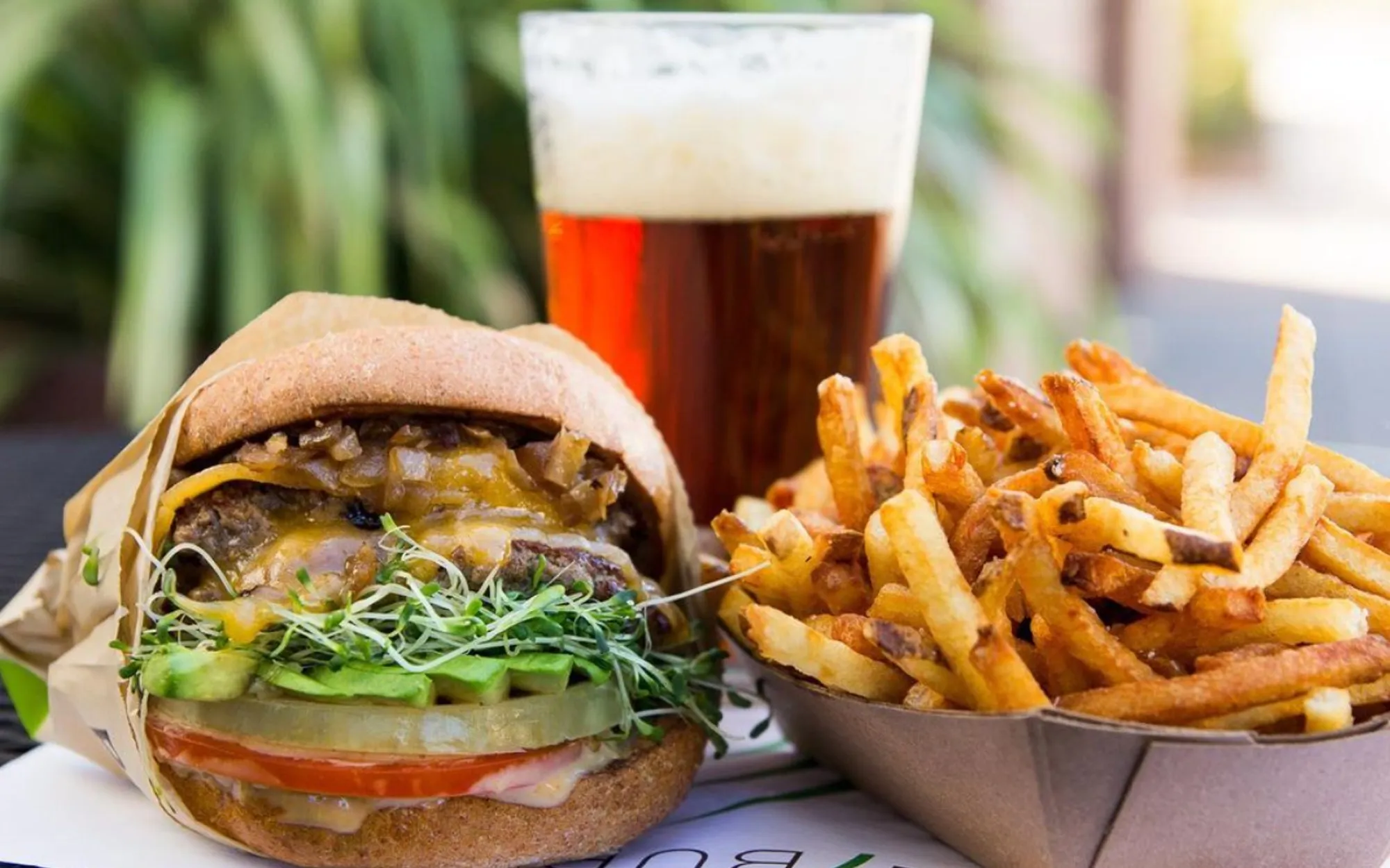 A juicy burger with melted cheese, onions, and sprouts, next to crispy fries in a tray, and a glass of frothy beer.