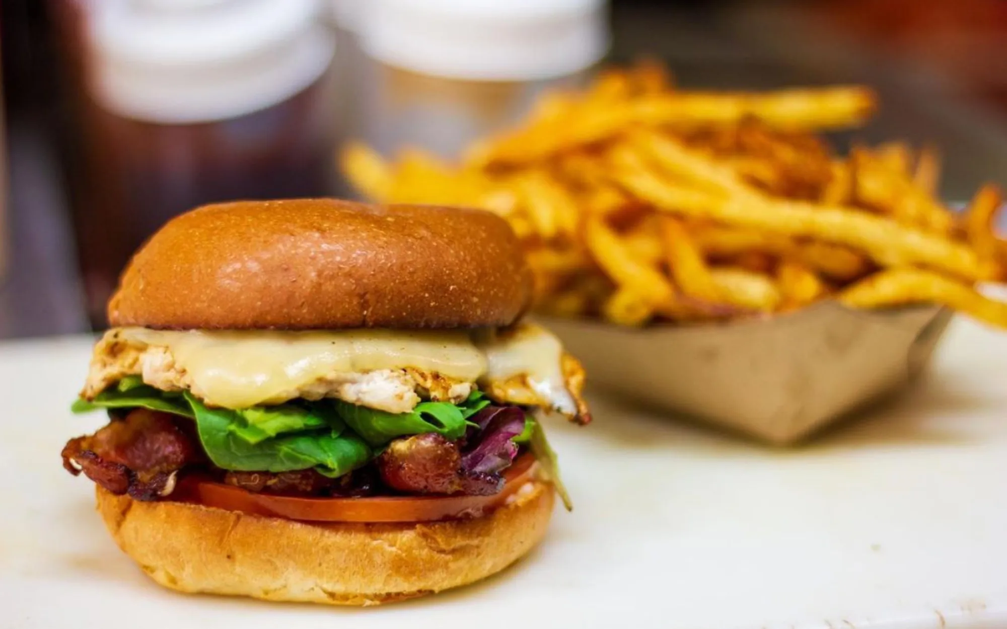 A juicy burger with bacon, cheese, greens, and tomato on a bun sits in the foreground. In the background, a tray of crispy fries adds appetizing warmth.
