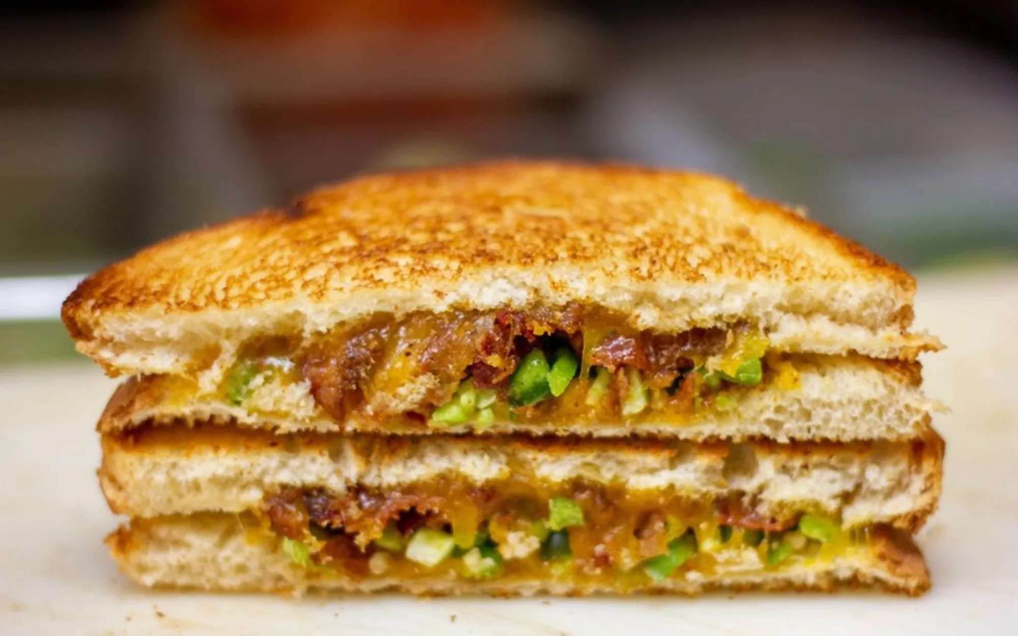 A toasted sandwich sliced in half, revealing layers of melted cheese, bright green peppers, and caramelized onions. The bread is golden brown and crispy.
