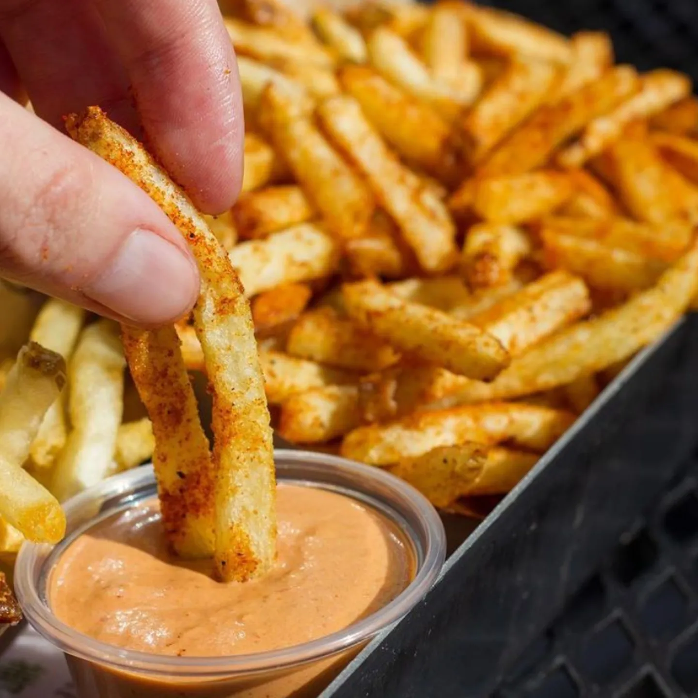 A hand dips seasoned fries into a small cup of creamy orange sauce. The fries are golden brown, suggesting crispiness.