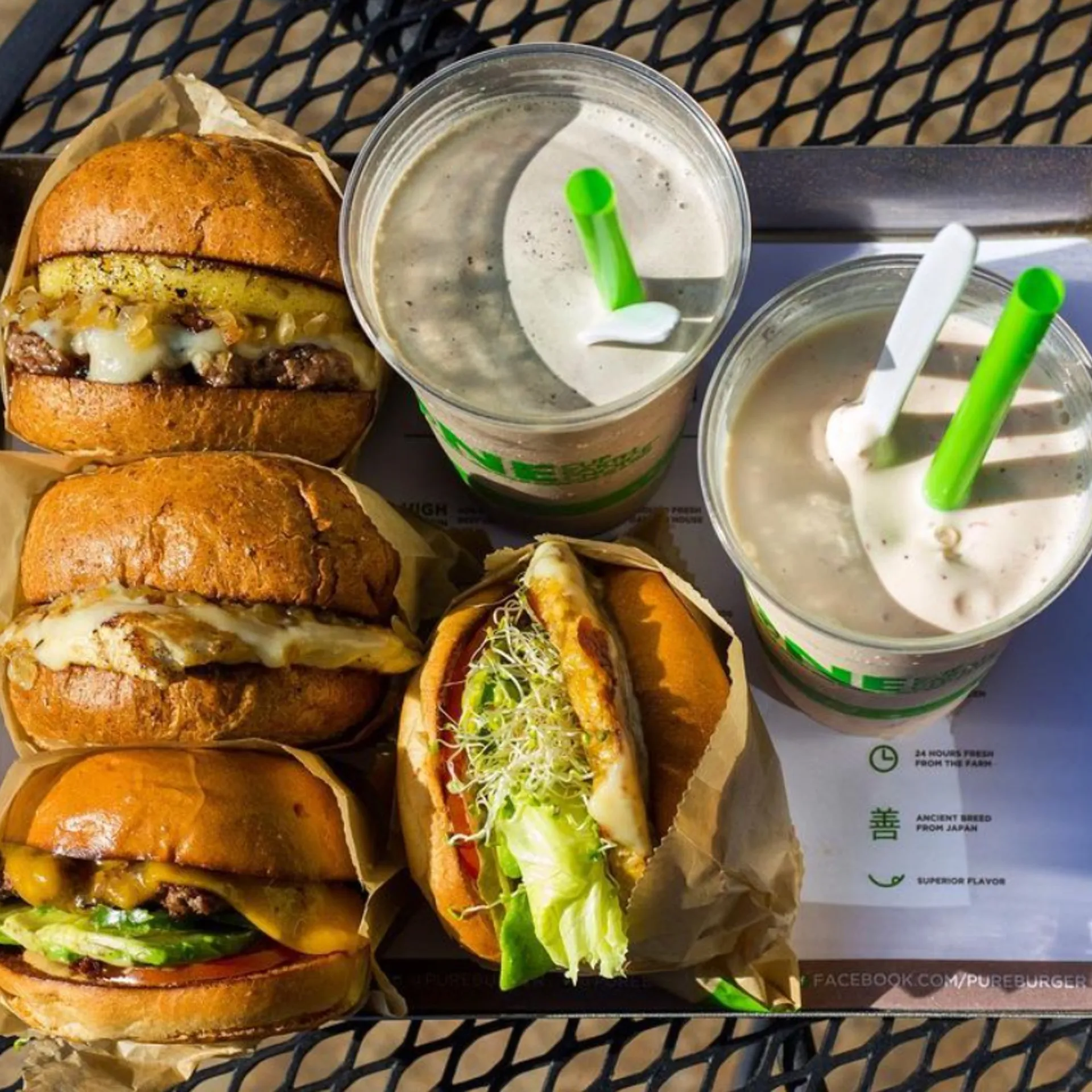 A tray holds four assorted burgers with golden buns and vibrant toppings, alongside two creamy milkshakes with green straws, set on a sunny day.
