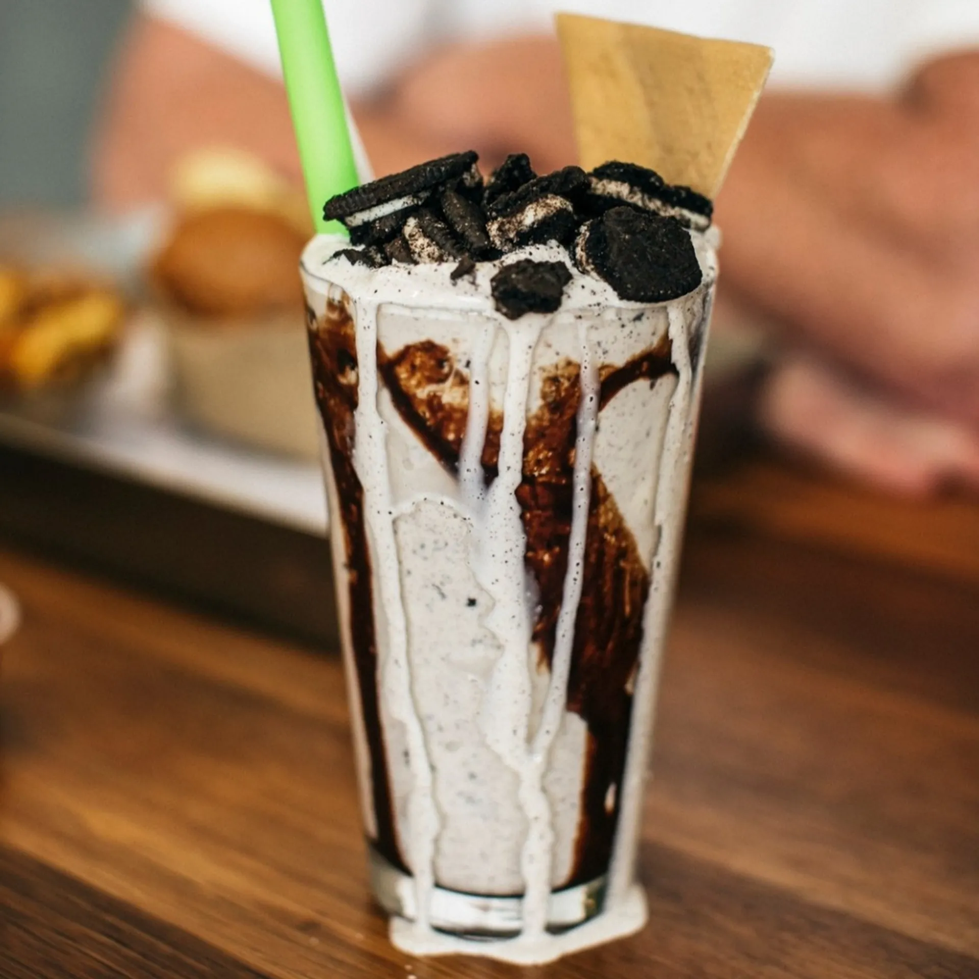A tall glass of cookies and cream milkshake with chocolate drizzle, crushed cookies, a green straw, and a wafer, sitting on a wooden table.