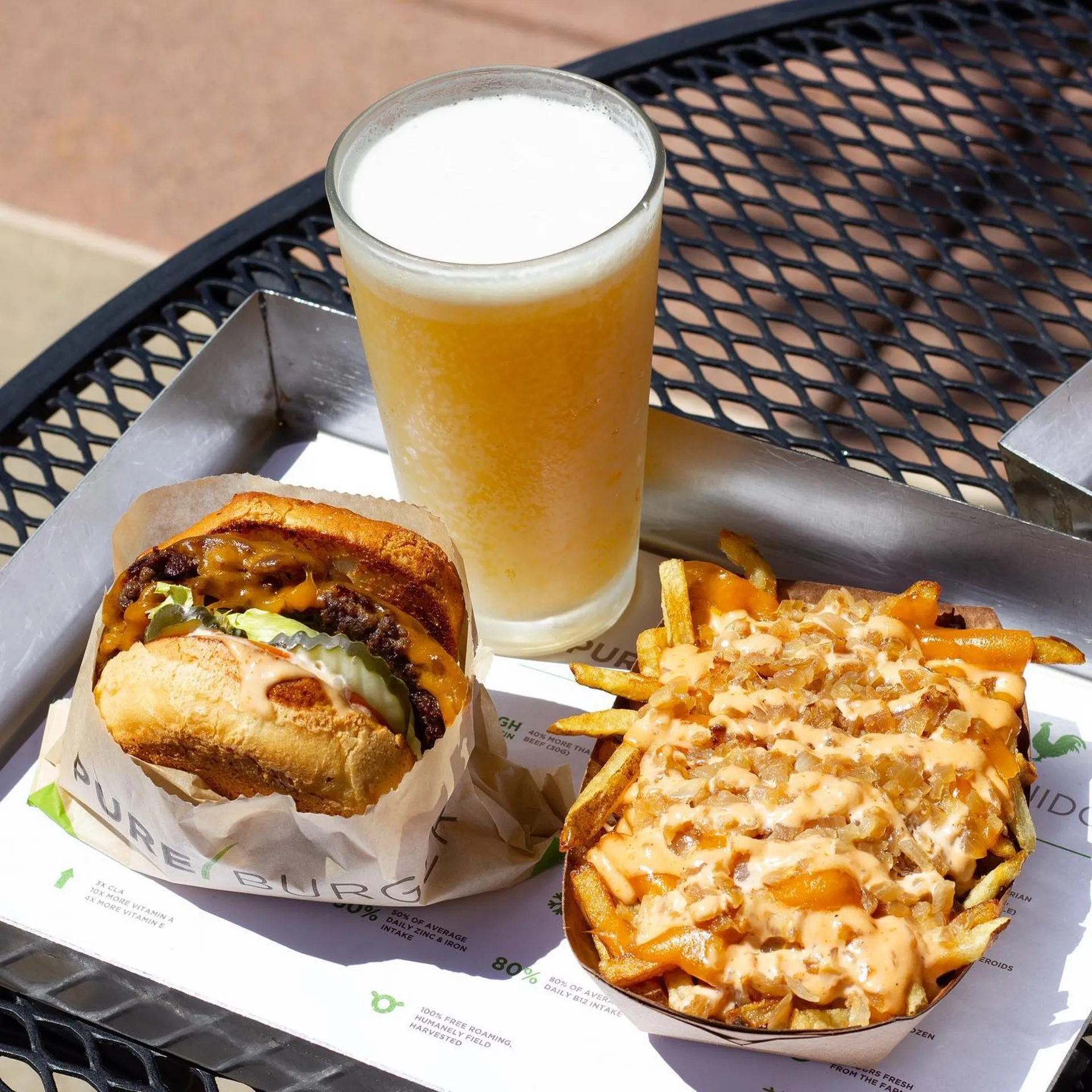 A juicy cheeseburger sits beside loaded fries drizzled with sauce in a metal tray. A frosty glass of beer completes the appetizing scene.