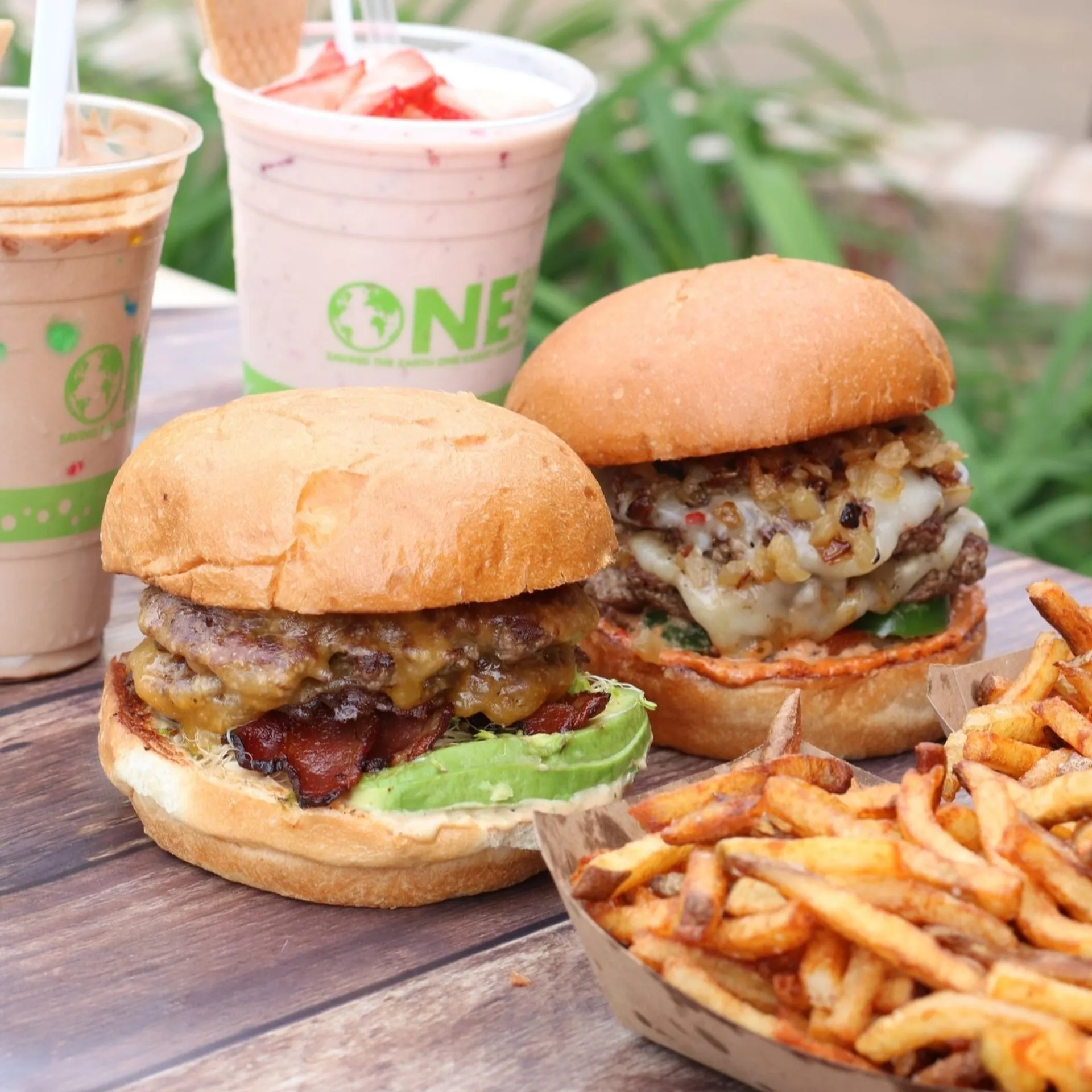Two burgers with melted cheese and bacon on a wooden table, accompanied by crispy fries. In the background, there are two milkshakes.
