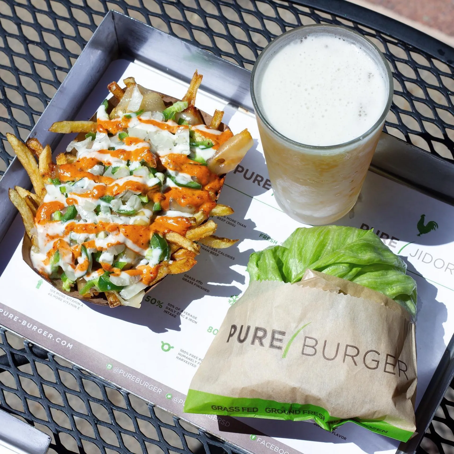 A tray with loaded fries drizzled with orange and white sauces, a lettuce-wrapped burger in branded paper, and a foamy drink on a wire table.