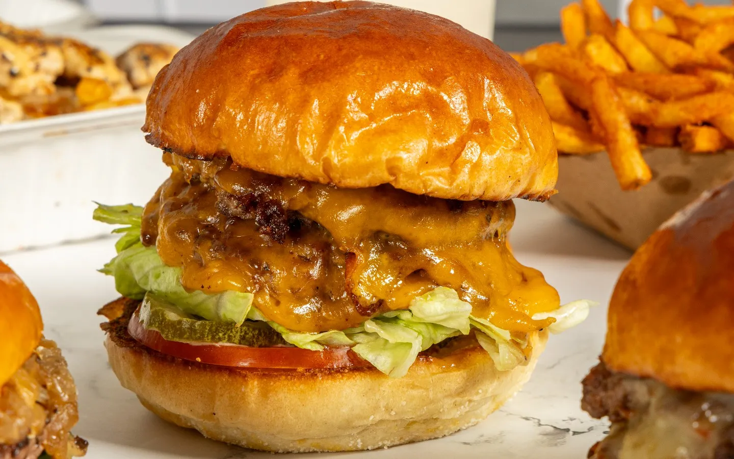 Close-up of a juicy cheeseburger with a glossy bun, lettuce, tomato, and pickles. Melted cheese oozes over the patty. Crispy fries are in the background.