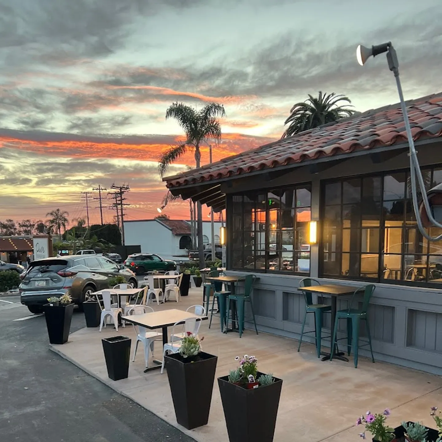 Outdoor seating at a restaurant with potted plants under a colorful sunset. Palm trees silhouette the sky, creating a serene, inviting atmosphere.