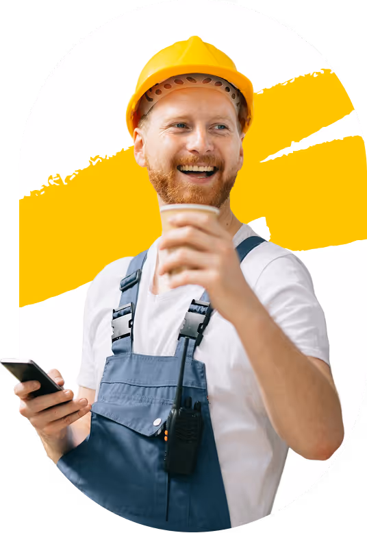 Ouvrier souriant avec casque de chantier jaune, salopette bleue, tenant un smartphone dans une main et un gobelet dans l'autre.