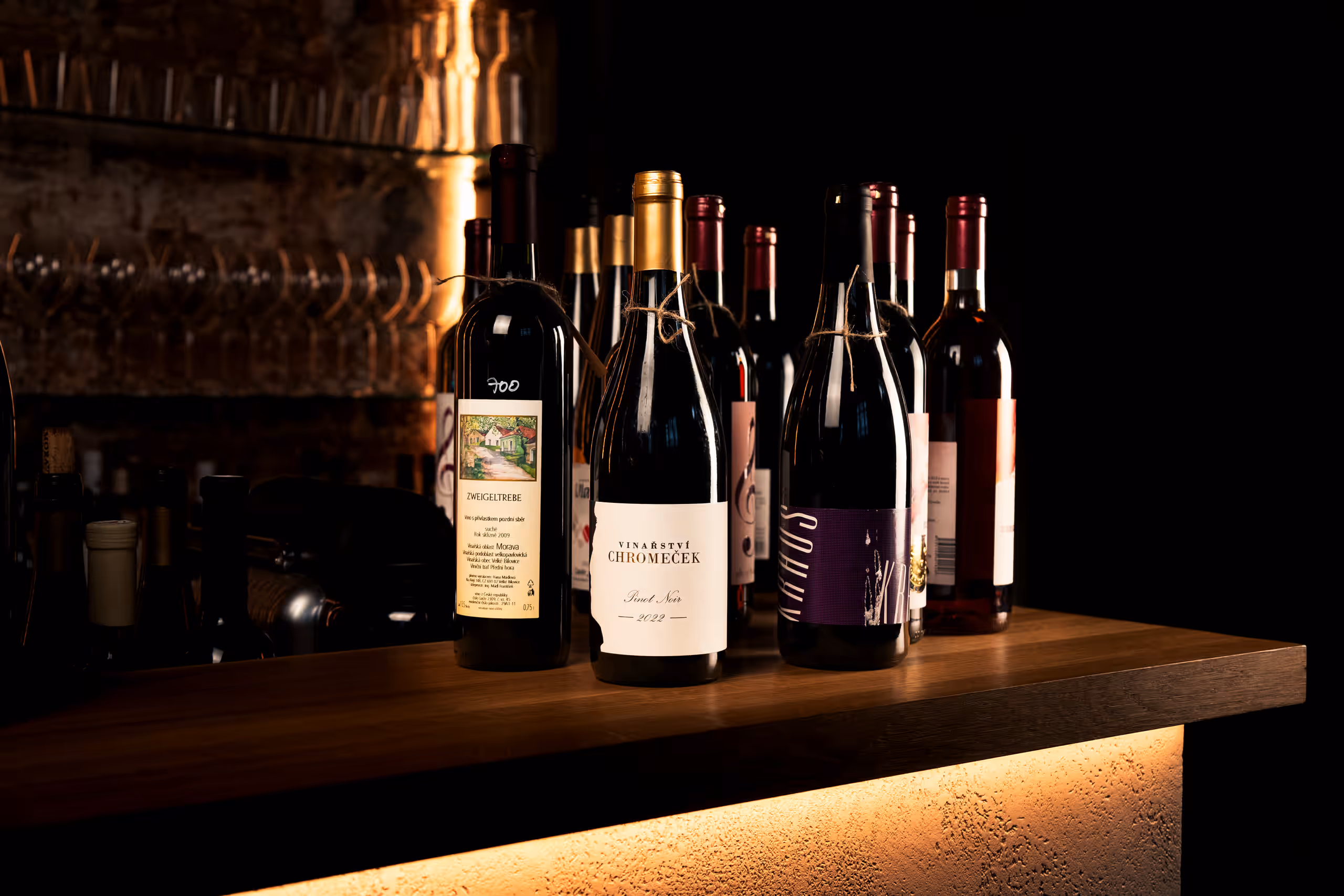 Několik lahví červeného vína seřazených na dřevěném pultu. Jedna láhev v popředí má etiketu "Vinařství Chromeček Pinot Noir 2022". Pozadí je tmavé s náznaky vinného sklepa nebo baru.