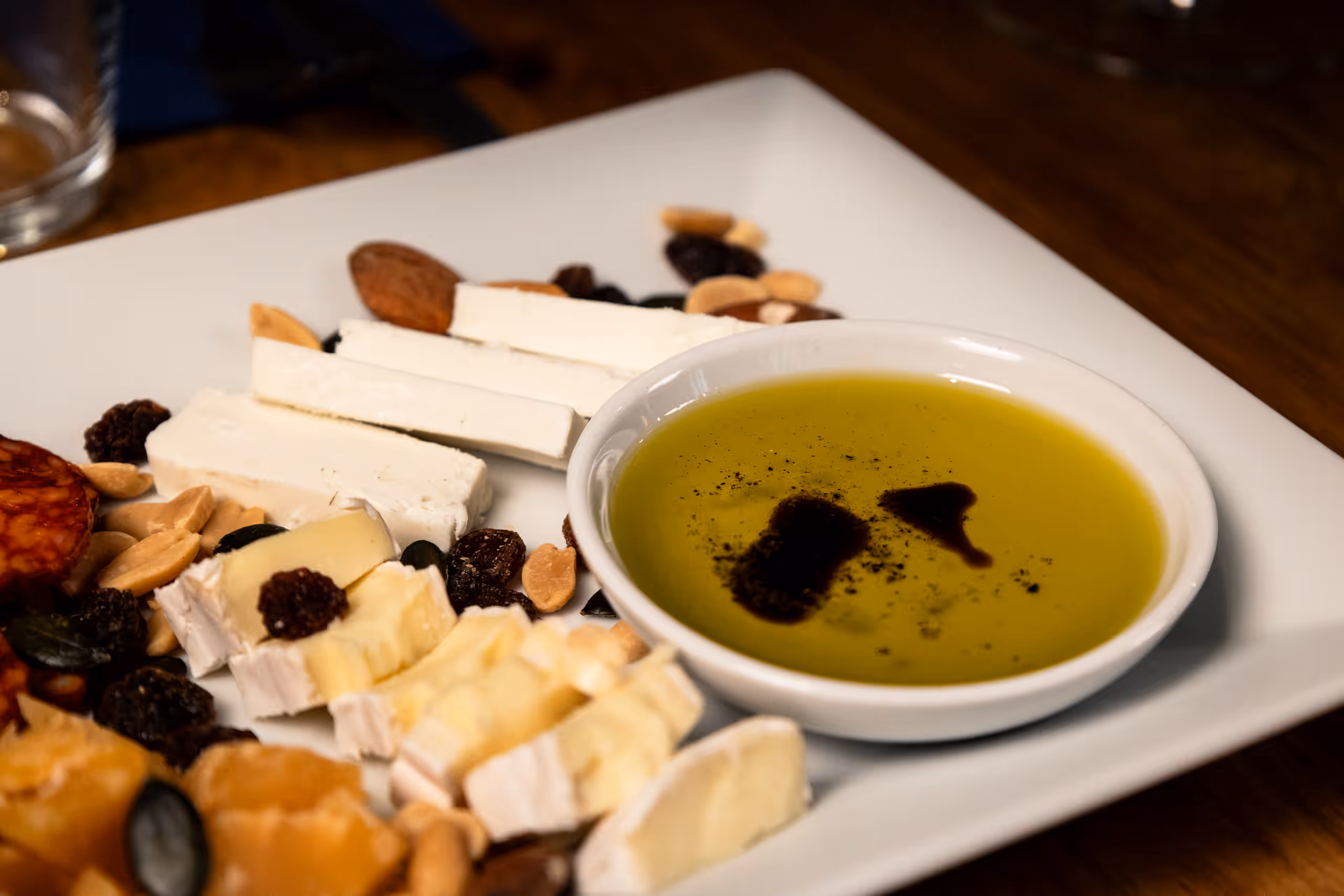 Detailní záběr na část obloženého prkénka, ukazující nakrájený bílý sýr, kousky měkkého sýra (jako brie nebo camembert) a malou bílou misku s olivovým olejem a balzamikovou glazurou. Kolem jsou rozmístěny ořechy a sušené ovoce.