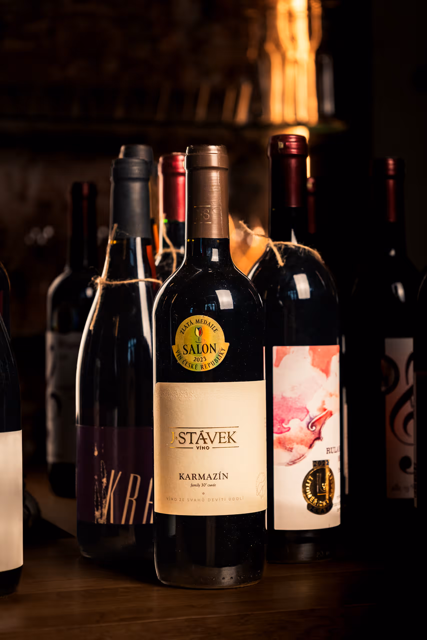Detailní záběr na láhev červeného vína s etiketou "J. Stávek Víno Karmazín family XO cuvée" a nálepkou "Zlatá medaile Salon Vín České republiky". Další lahve vína jsou viditelné v rozostřeném pozadí.