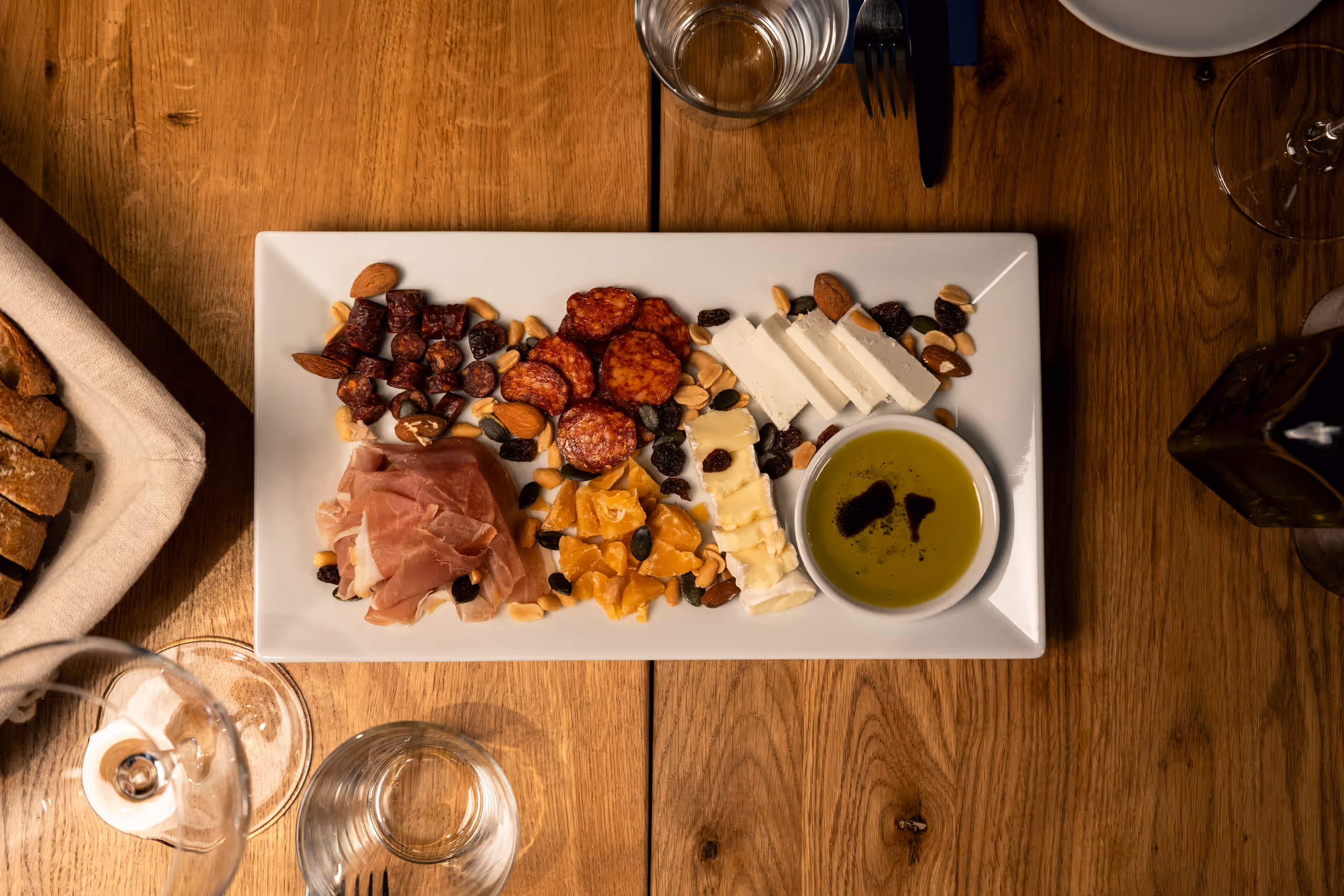 Obložené prkénko na dřevěném stole, pohled shora. Obsahuje různé druhy uzenin, sýrů, ořechů, sušeného ovoce a malou misku s olivovým olejem.