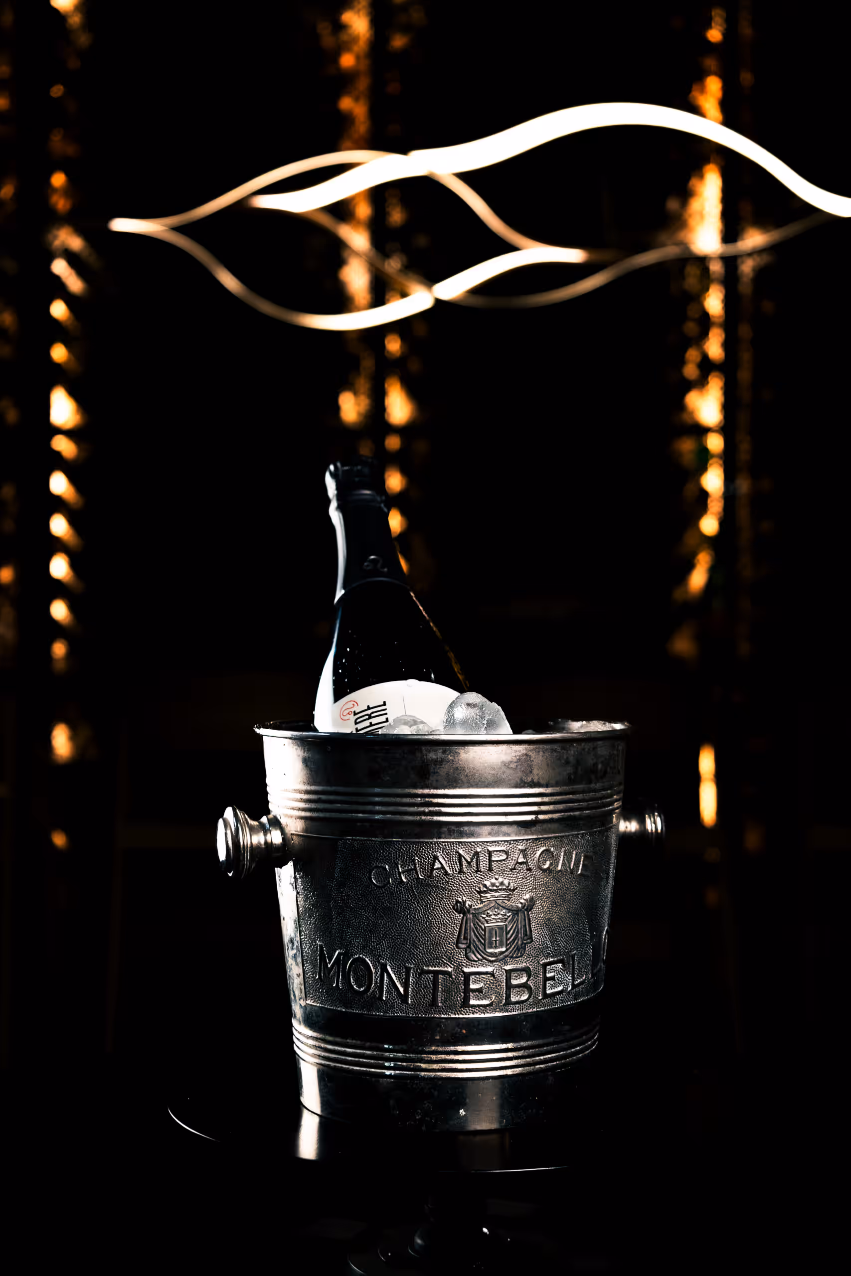 Láhev šumivého vína ve stříbrném kbelíku na šampaňské s nápisem "CHAMPAGNE MONTEBELLO". Pozadí je tmavé s abstraktními světelnými pruhy.