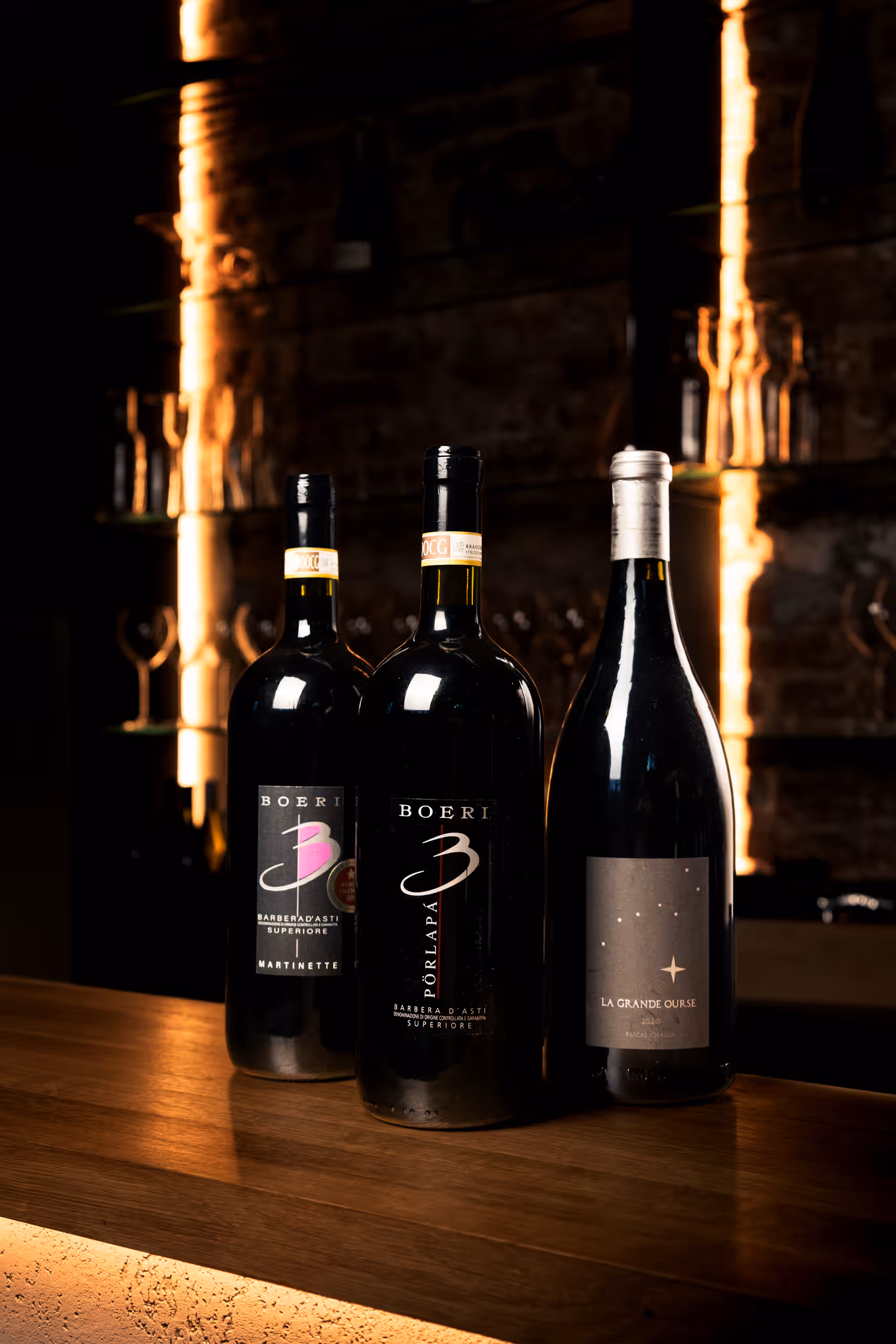Tři lahve červeného vína na dřevěném pultu. Etikety ukazují "Boeri Barbera d'Asti Superiore Martinette", "Boeri Porlapà Barbera d'Asti Superiore" a "La Grande Ourse". Pozadí tvoří tlumeně osvětlený bar nebo sklep.