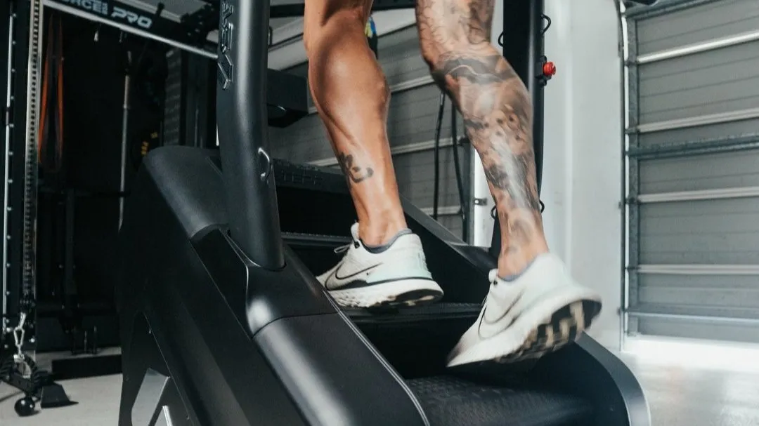 Close-up of a person with tattoos wearing white Nike sneakers using a stair-stepper machine indoors.
