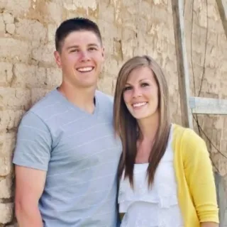 Smiling young couple standing close together in front of a weathered brick wall.