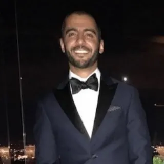 Smiling man wearing a dark tuxedo with a black bow tie standing against a nighttime cityscape background.