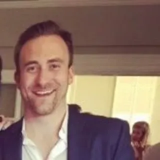 Smiling man with short brown hair and a trimmed beard wearing a dark blazer over a white shirt indoors.