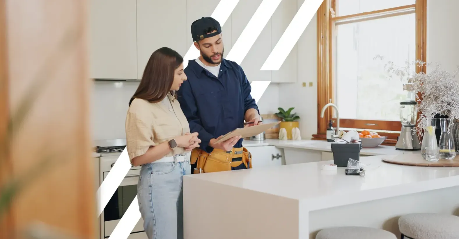 Un technicien en bleu avec une ceinture à outils montre un document sur une planchette à une femme dans une cuisine moderne.