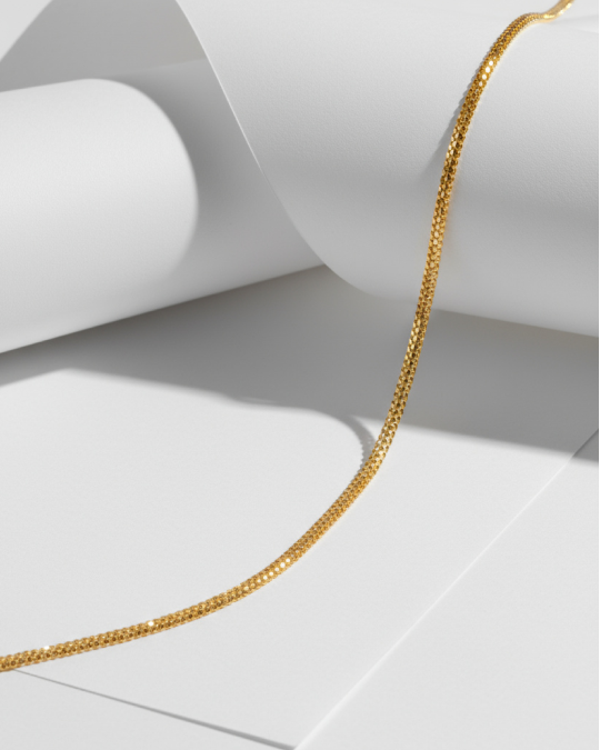 Delicate gold chain necklace draped over curved white paper sheets casting soft shadows.